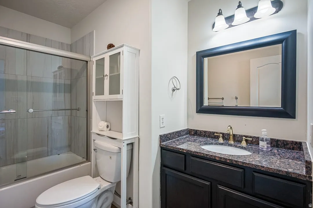 Bathroom featuring a dark wood vanity with a stone countertop, a framed mirror, a three-light vanity fixture, a toilet, and a tub with a sliding glass door and tiled surround