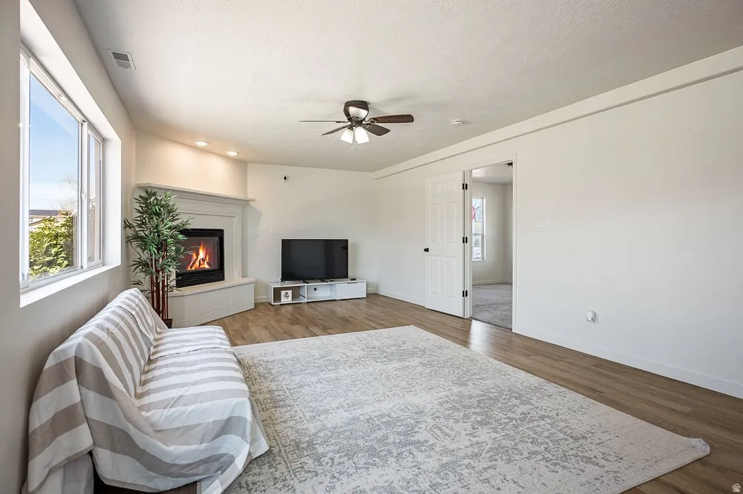 Open living area featuring wood-finish flooring, a corner fireplace with a white mantel, and large windows