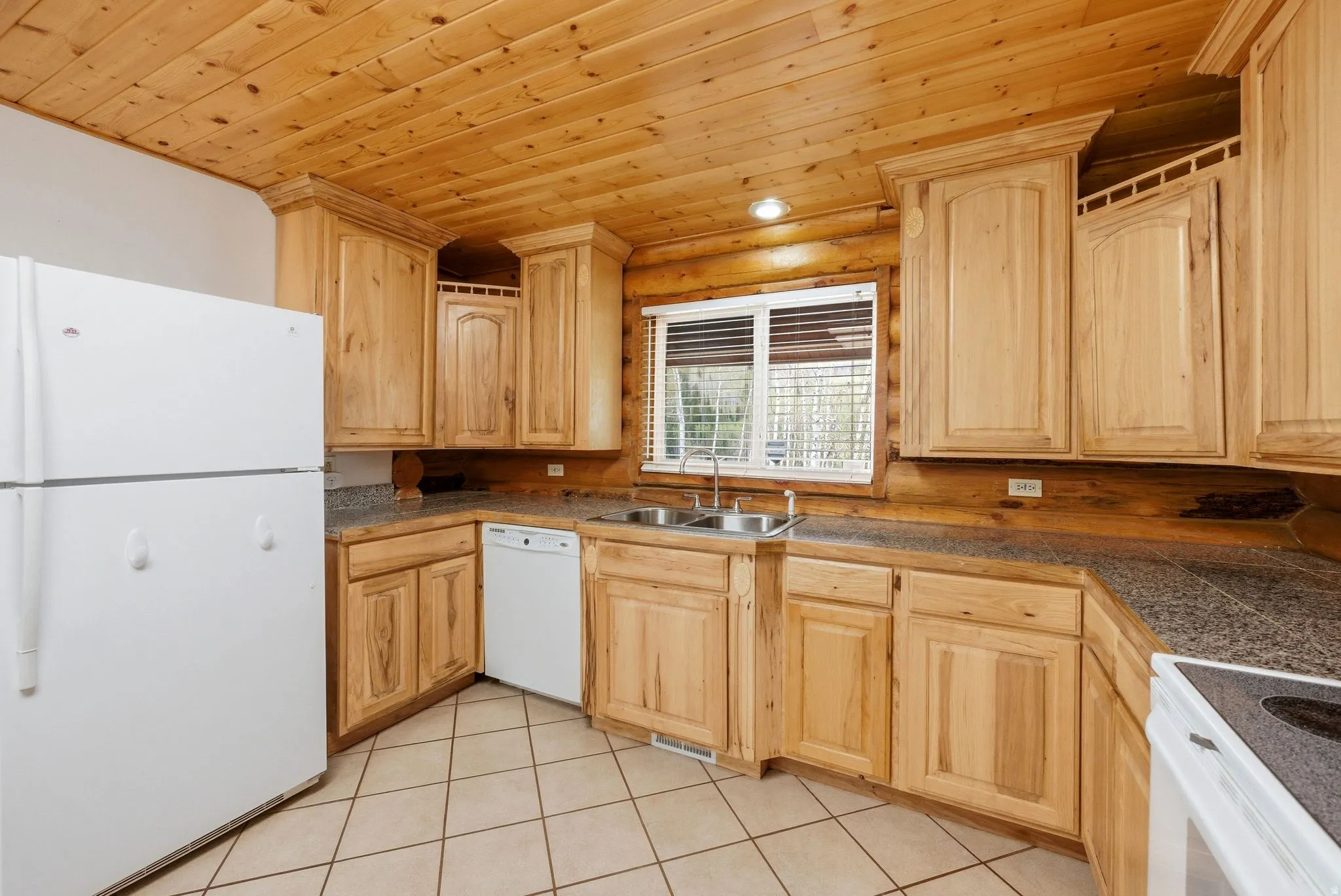 Kitchen featuring natural wood cabinetry, wood-paneled ceiling, light-toned tile flooring, L-shaped countertop, and a double basin stainless steel sink