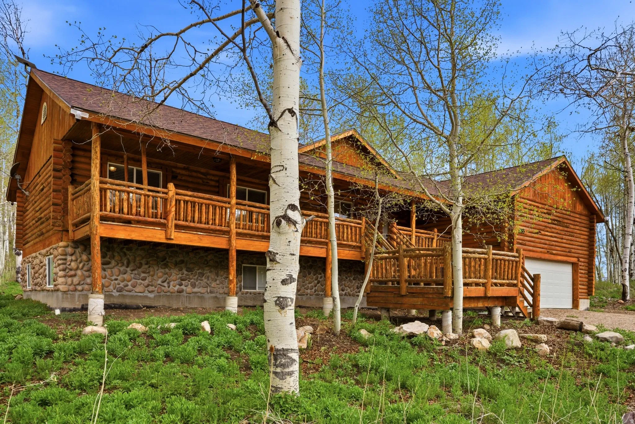 Log cabin exterior featuring a full-width covered porch, a stone foundation, a multi-level deck, and an integrated garage with a white door