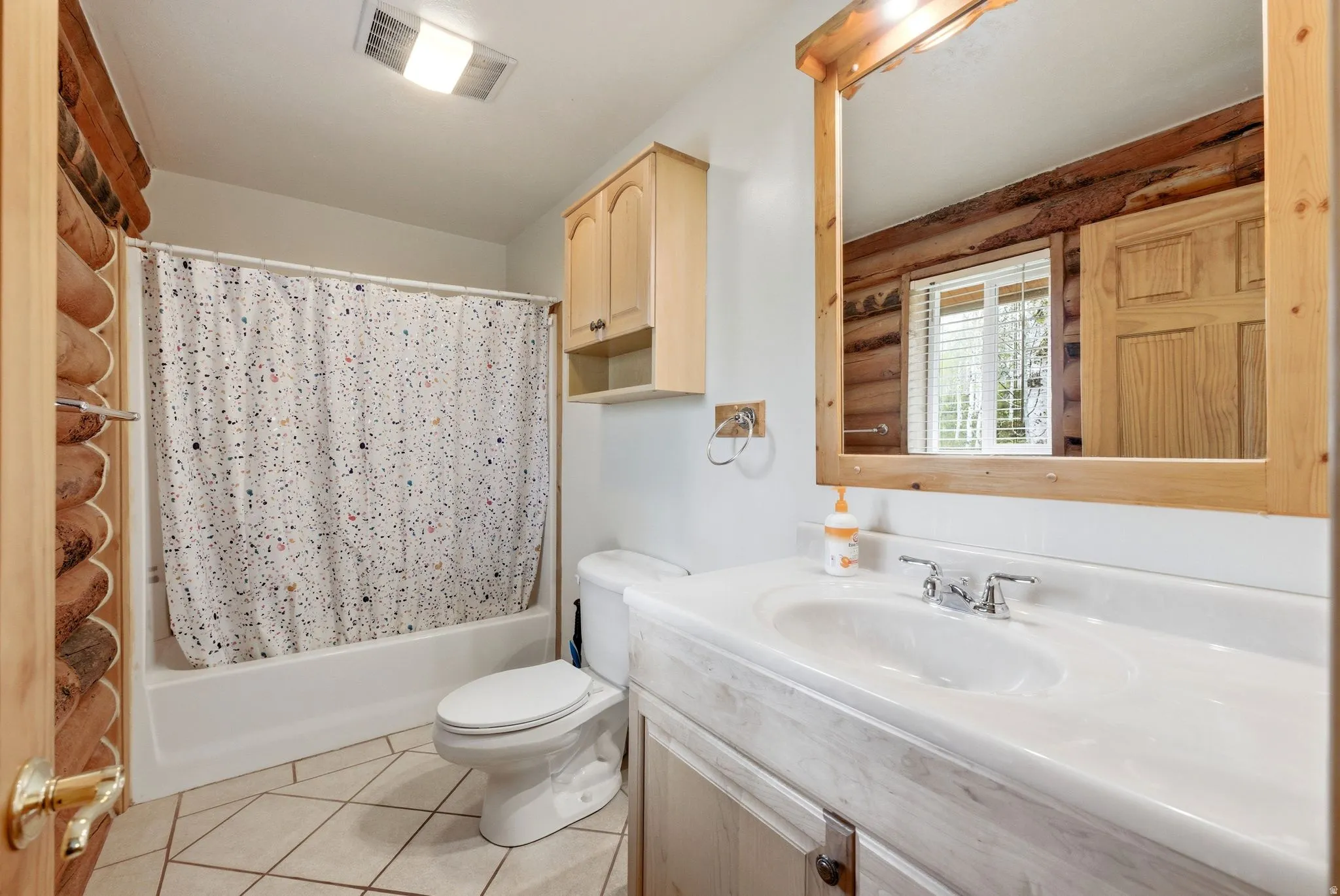Bathroom featuring log wall construction, a single-sink vanity with a white countertop, a mirror with a wood frame, and light-toned tile flooring
