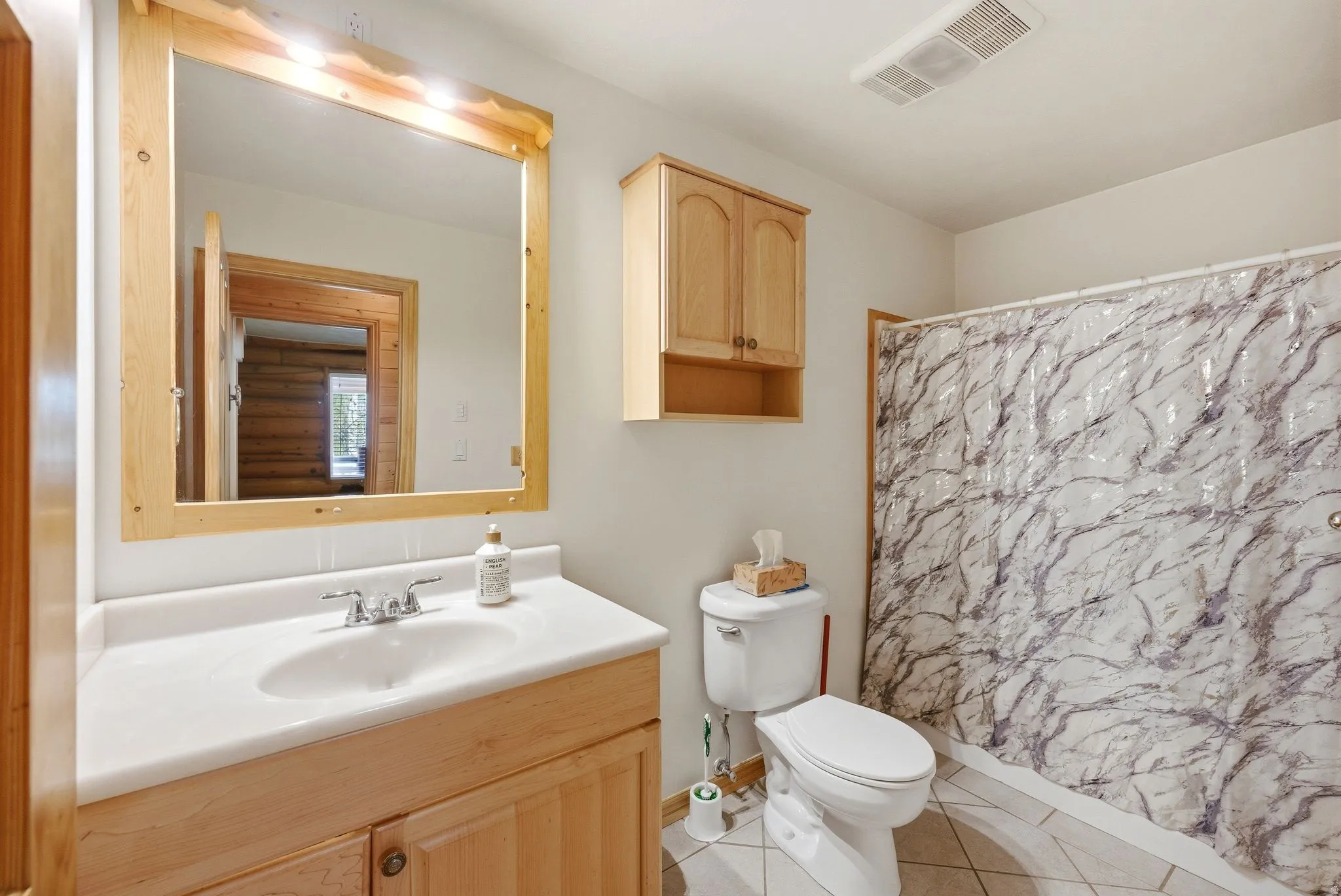 Bathroom featuring a vanity with an integrated sink, a wood-frame mirror with overhead lighting, a toilet, a shower area, and a wall-mounted wood cabinet