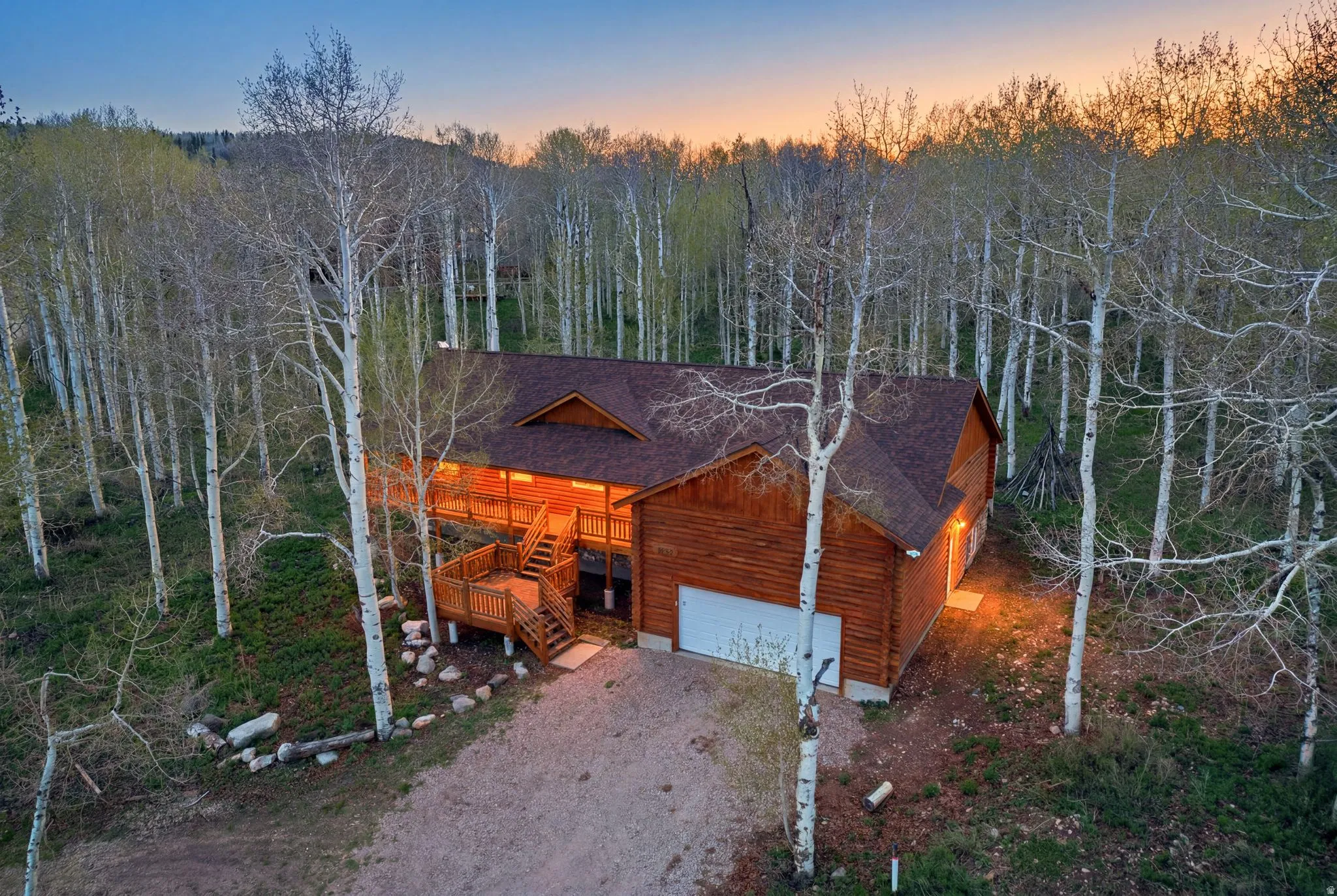 Log-sided residence surrounded by mature trees, featuring a prominent front deck with stairs, a multi-level roofline, an attached garage with a white door, and a gravel driveway