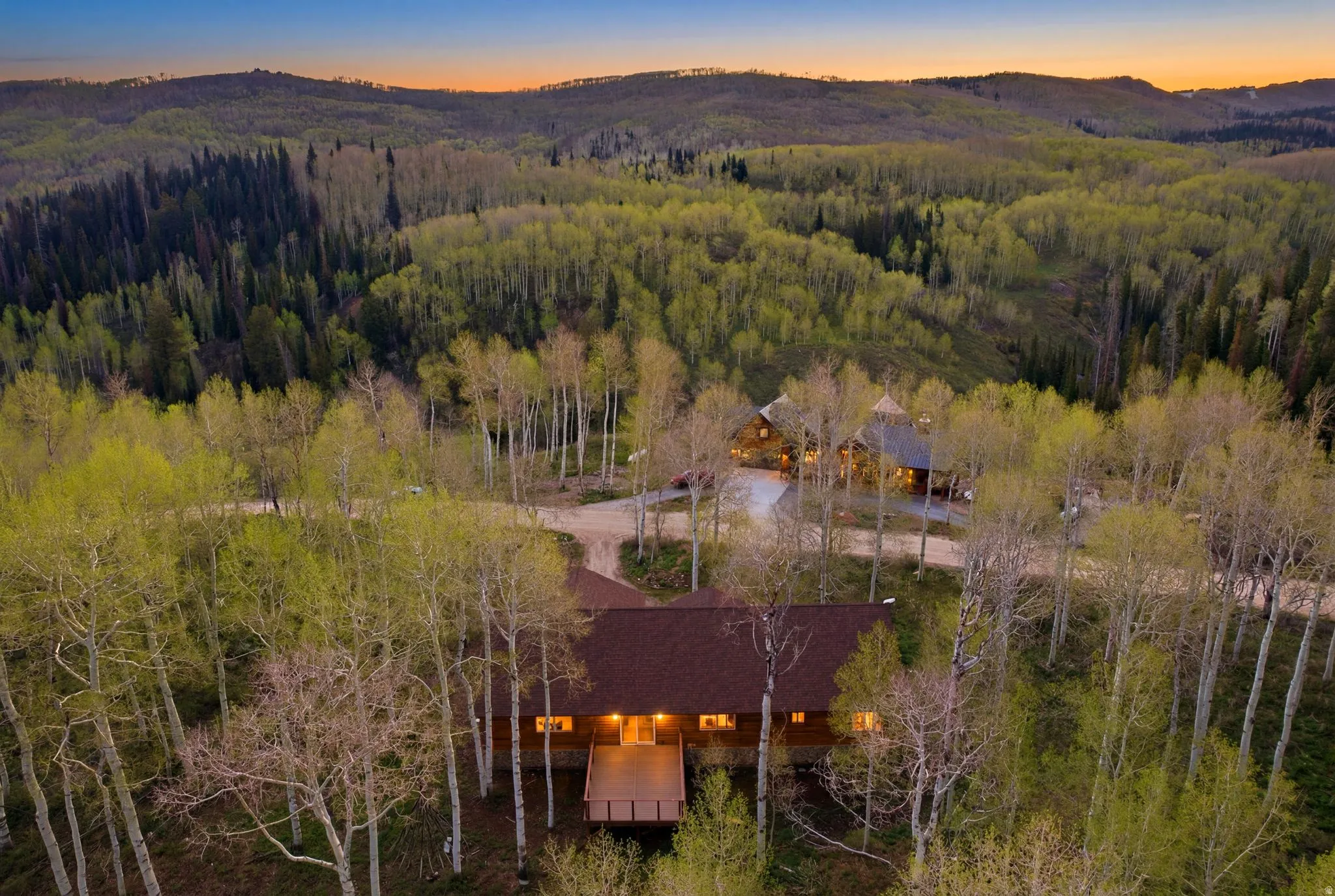 Wood-clad residence featuring a spacious deck and illuminated windows, surrounded by abundant mature trees