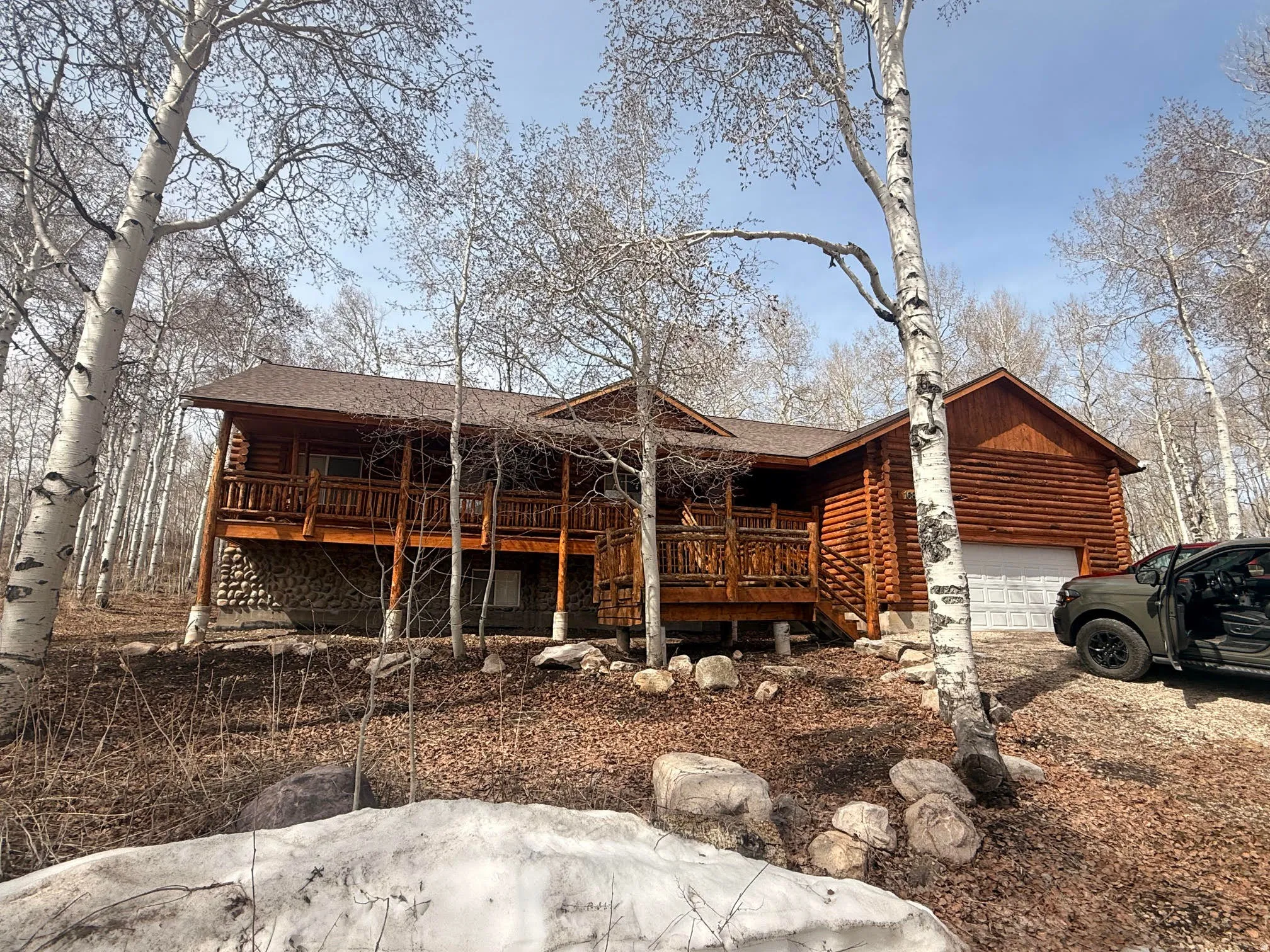 Log cabin exterior featuring a covered porch, stone foundation, attached garage, and surrounding mature trees