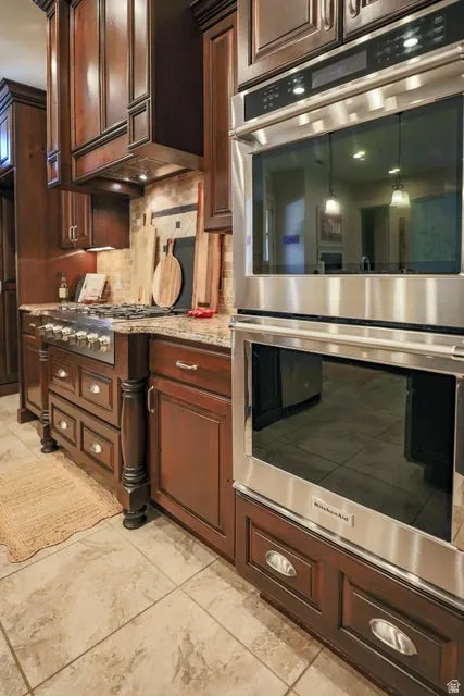 Gourmet kitchen featuring a stainless steel double wall oven, a professional-grade gas range, custom wood cabinetry with decorative corbels, stone-finish countertops, and a tiled backsplash