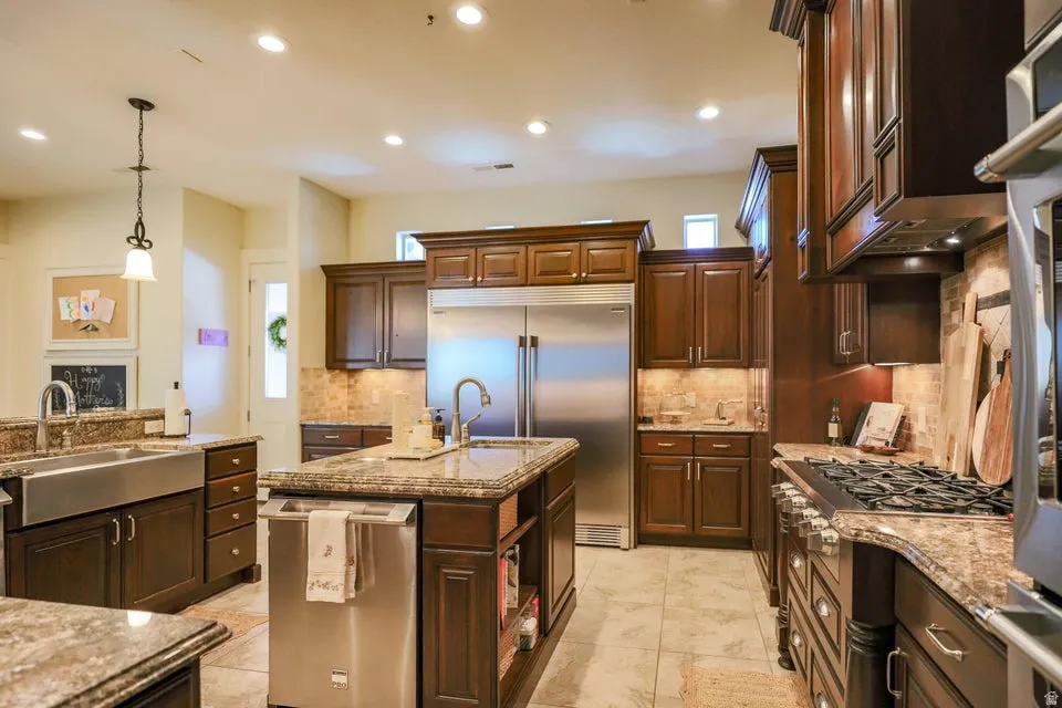 Kitchen featuring rich wood cabinetry, stone countertops, stainless steel appliances, a center island with sink, and tile flooring