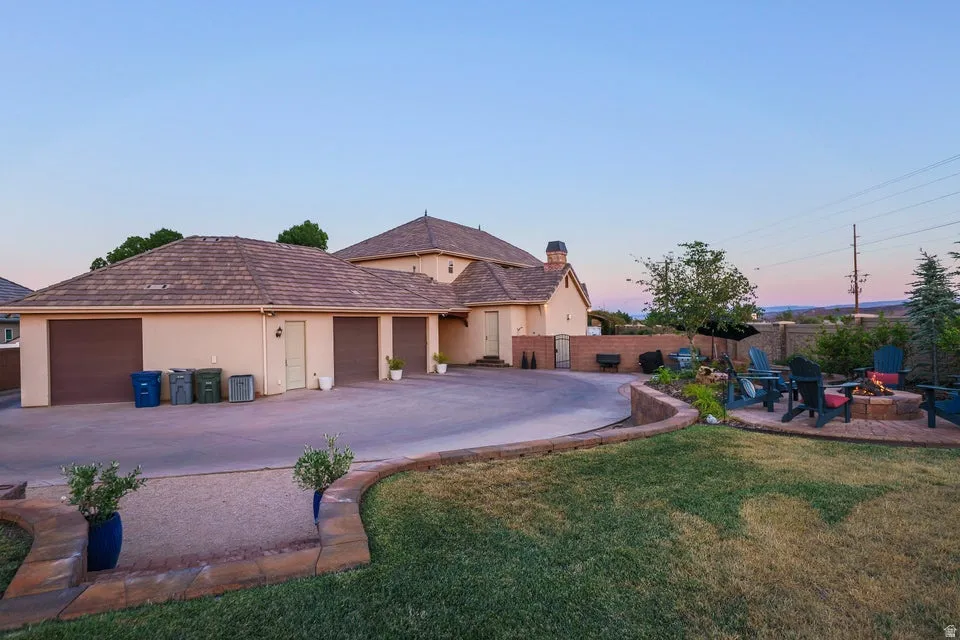 Exterior stucco residence with a multi-car garage, featuring a curved driveway, a retaining wall, a manicured lawn, and an outdoor fire pit area
