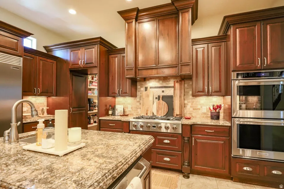 Gourmet kitchen featuring rich wood cabinetry, stainless steel appliances including a double oven and gas range, mosaic tile backsplash, and stone countertops