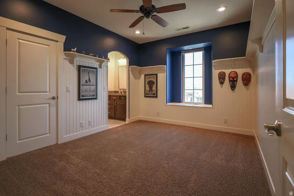 Carpeted room featuring a white paneled wainscoting and deep blue walls, a multi-pane window with a deep sill, a ceiling fan with integrated lighting, and an arched doorway leading to a vanity area