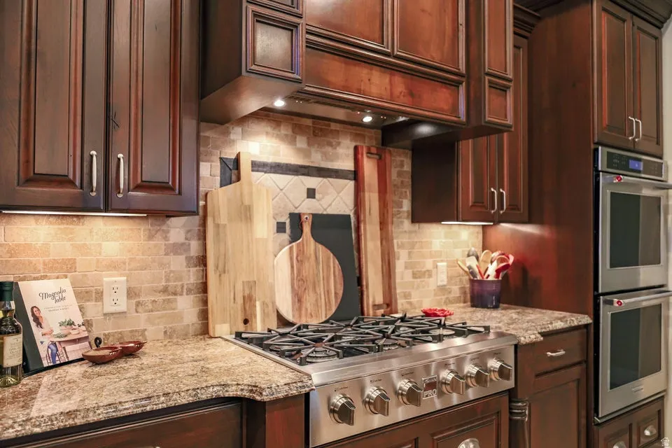 Gourmet kitchen featuring a stainless steel gas range, double wall ovens, granite countertops, stone tile backsplash, and rich wood cabinetry