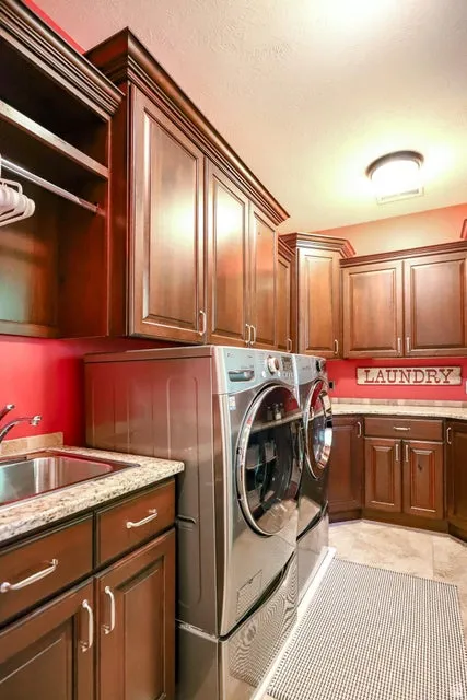 Laundry room featuring extensive dark wood cabinetry with crown molding, a built-in stainless steel sink, light granite-finish countertops, and tiled flooring