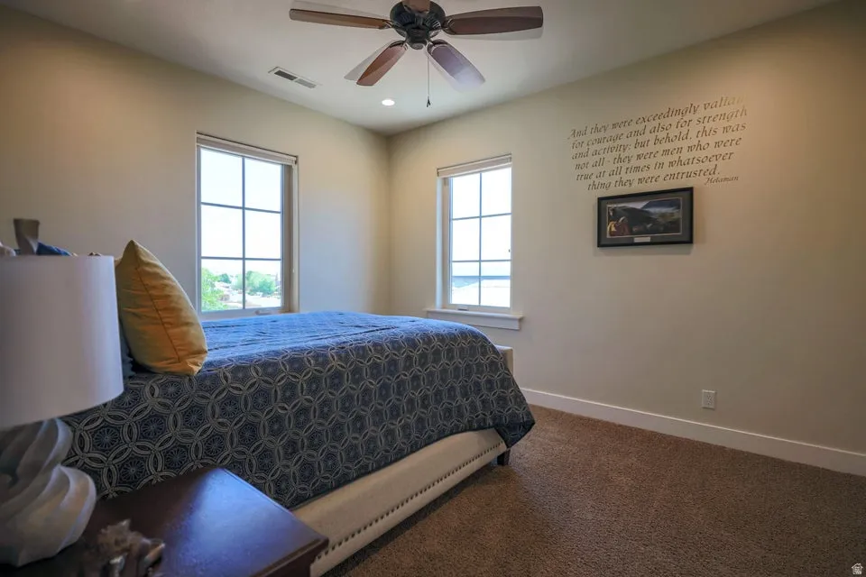 Carpeted bedroom featuring two large windows with exterior views and a ceiling fan with integrated lighting