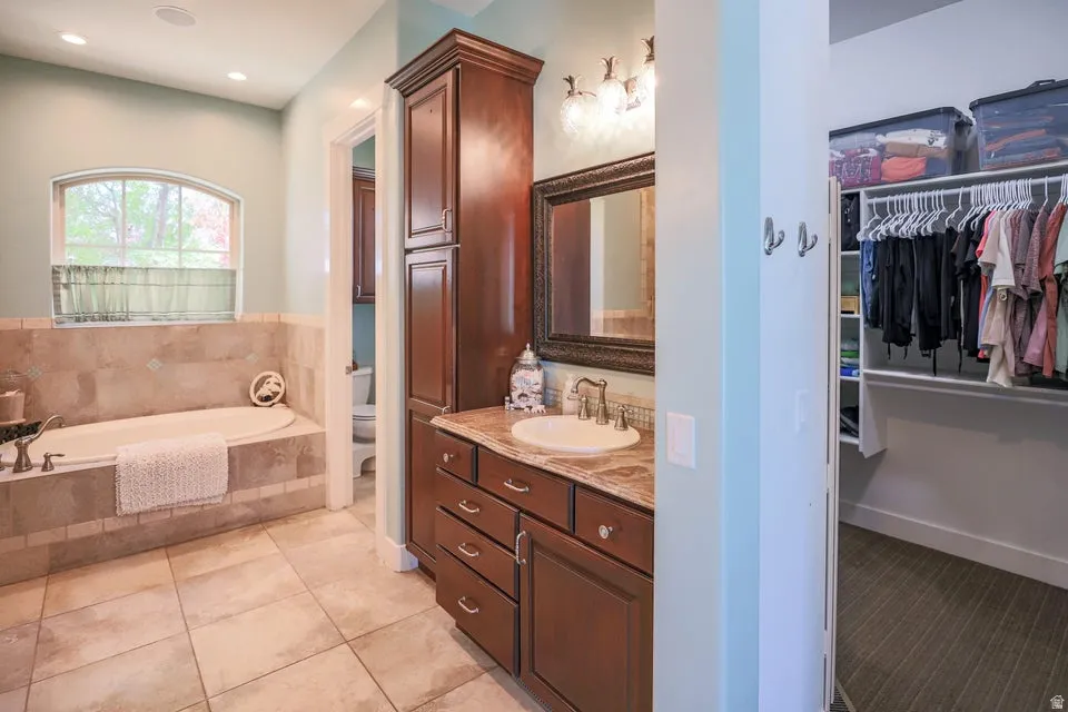 Deep soaking tub with tile surround, arched window, wood-finish vanity with countertop and integrated sink, and recessed lighting