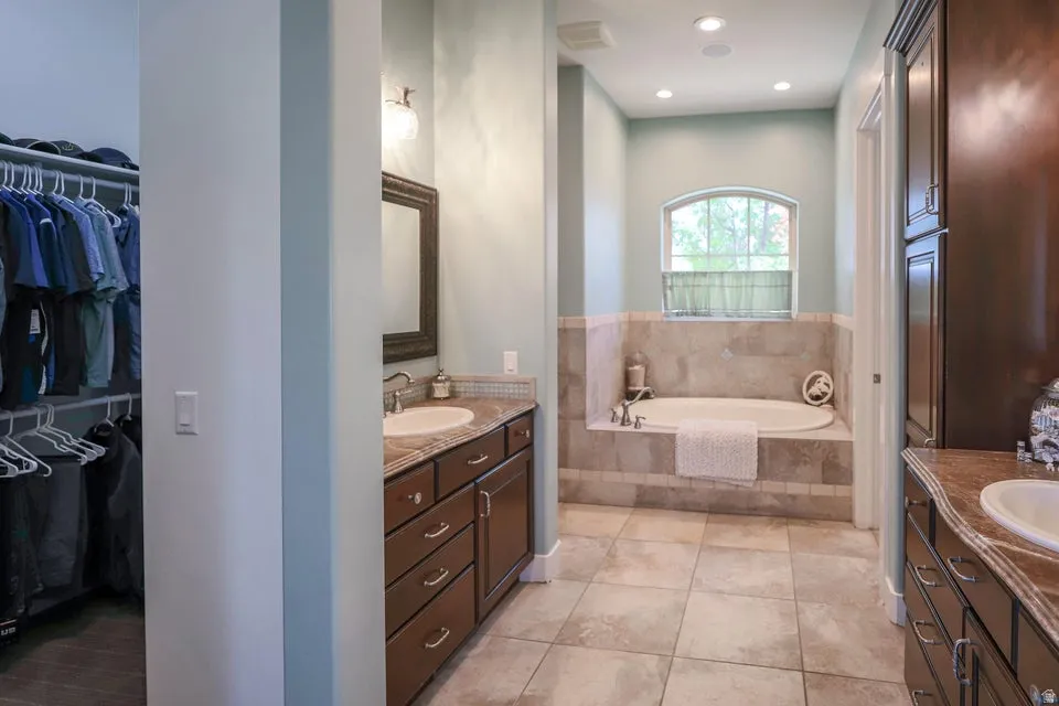 Spacious bathroom featuring a built-in soaking tub with a tiled surround, arched window, wood-finish cabinetry, and tiled flooring