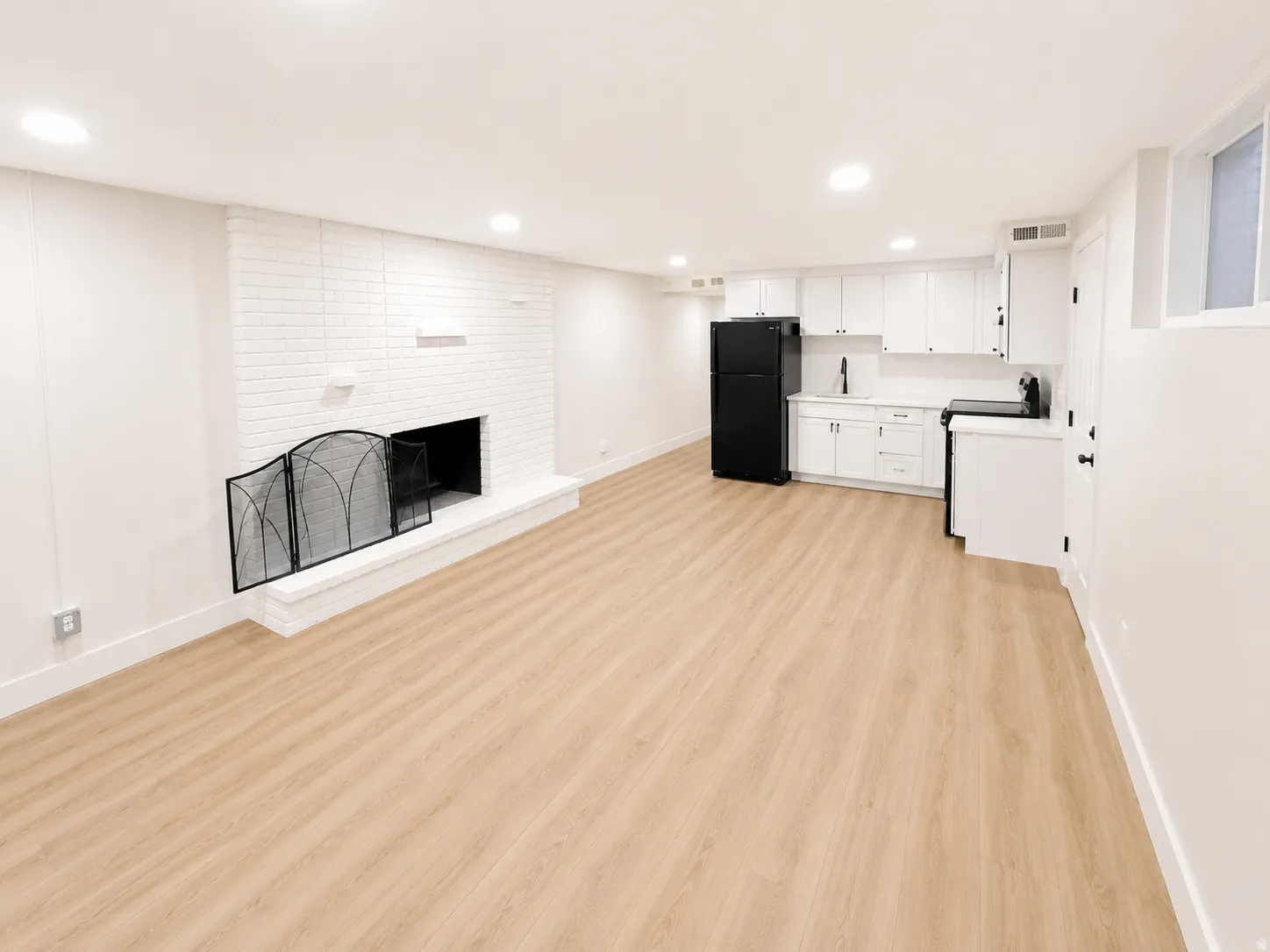 Open-concept space featuring a white brick fireplace with hearth, recessed lighting, wood-finish flooring, a full kitchen with white cabinetry and black appliances, and white walls