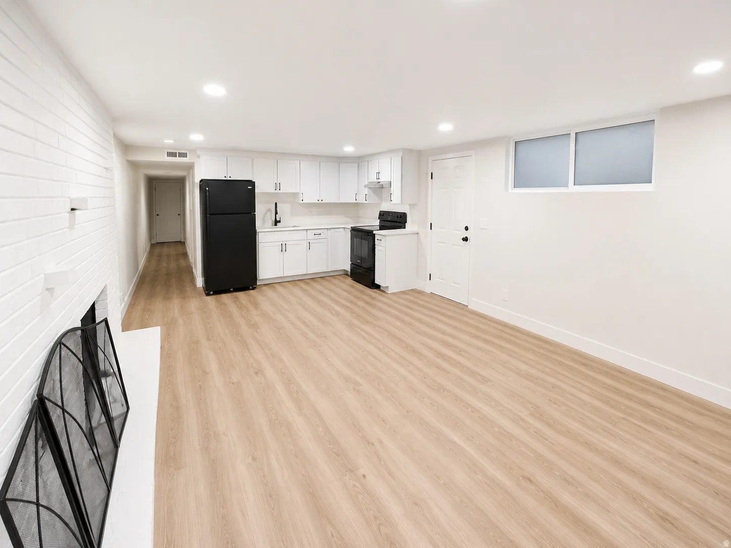 Open-concept living space featuring wood-finish flooring, a contemporary kitchen with white cabinetry and black appliances, recessed lighting, and a white brick-textured wall with an integrated fireplace