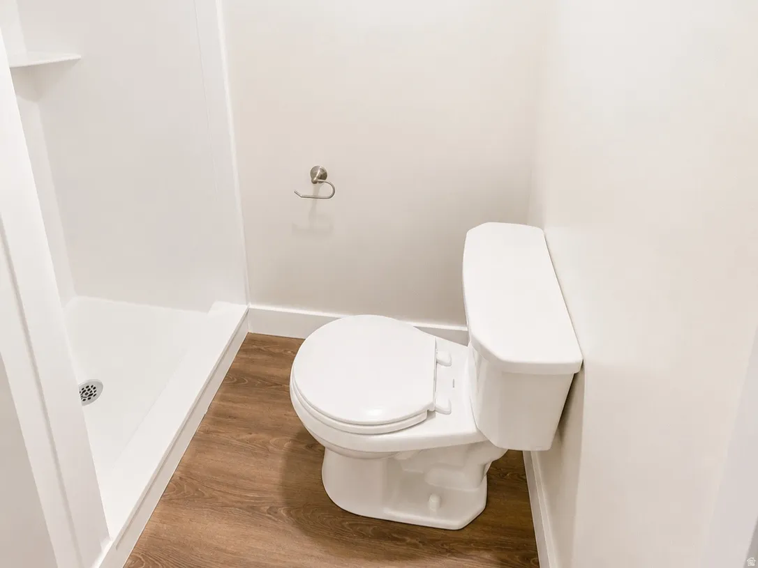 Full bathroom featuring a white toilet, enclosed shower stall with built-in shelving, wood-finish flooring, a privacy window, and a brushed nickel towel bar