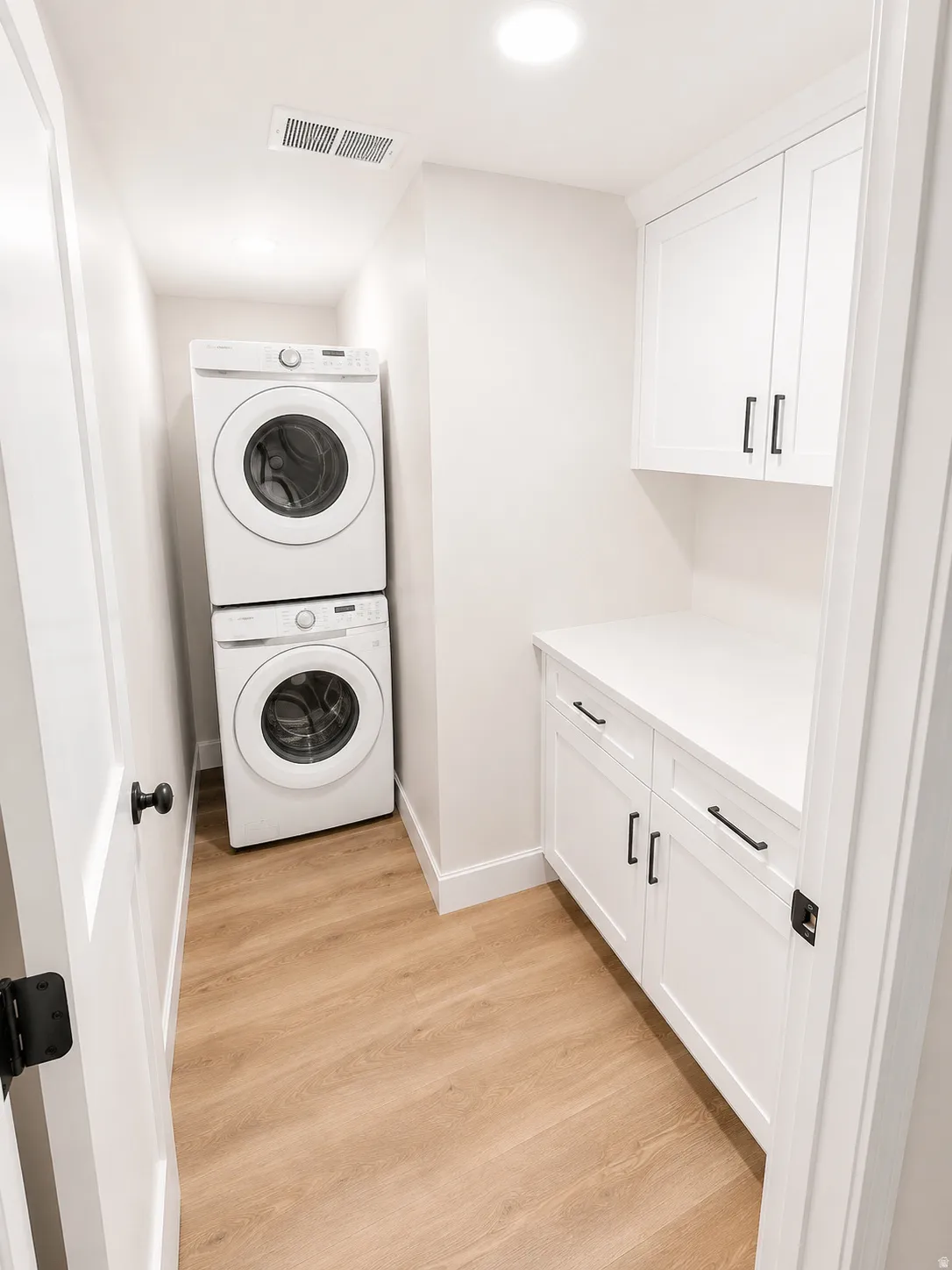 Dedicated laundry area featuring stacked appliances, wood-finish flooring, white cabinetry with black hardware, and a white countertop