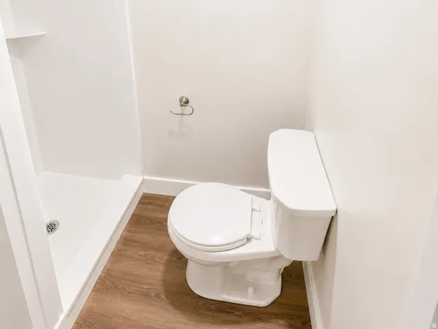 Full bathroom featuring a white toilet, enclosed shower stall with built-in shelving, wood-finish flooring, a privacy window, and a brushed nickel towel bar