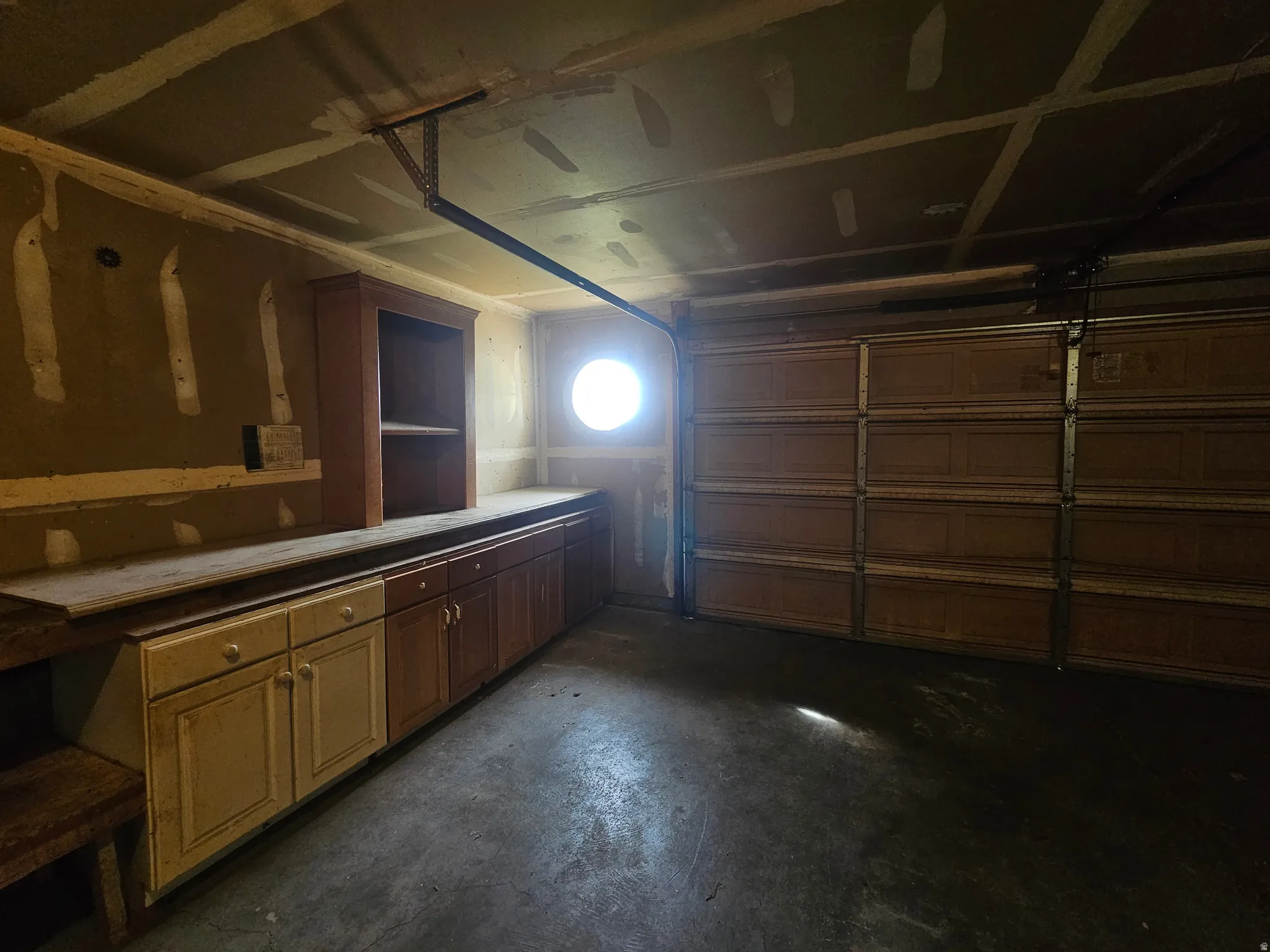 Garage interior featuring a built-in workbench with storage cabinetry, an overhead garage door, a circular wall-mounted window, exposed ceiling joists, and a concrete floor