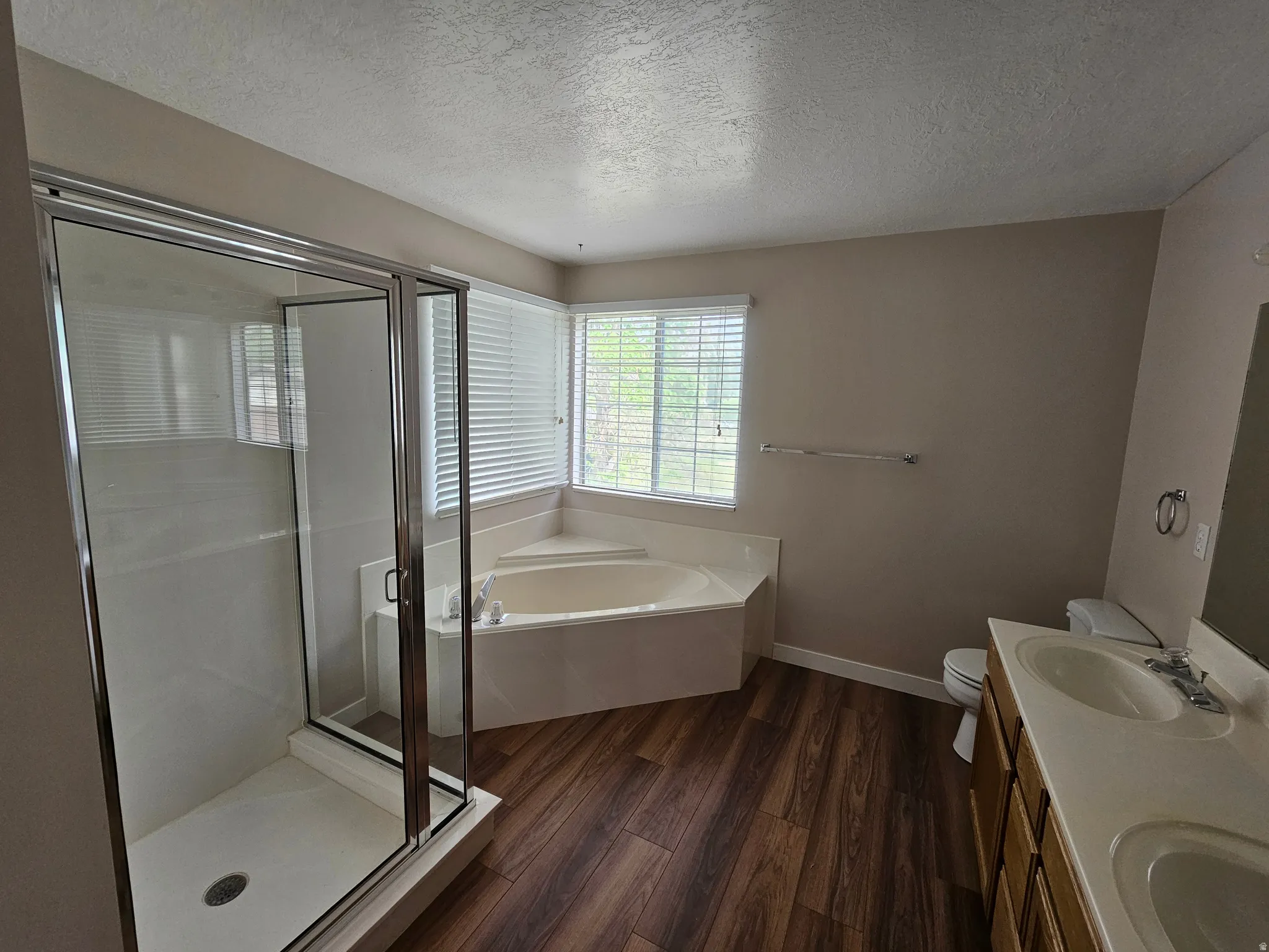 Bathroom featuring a glass-enclosed shower, corner soaking tub, dual-sink vanity, wood-finish flooring, and horizontal window blinds