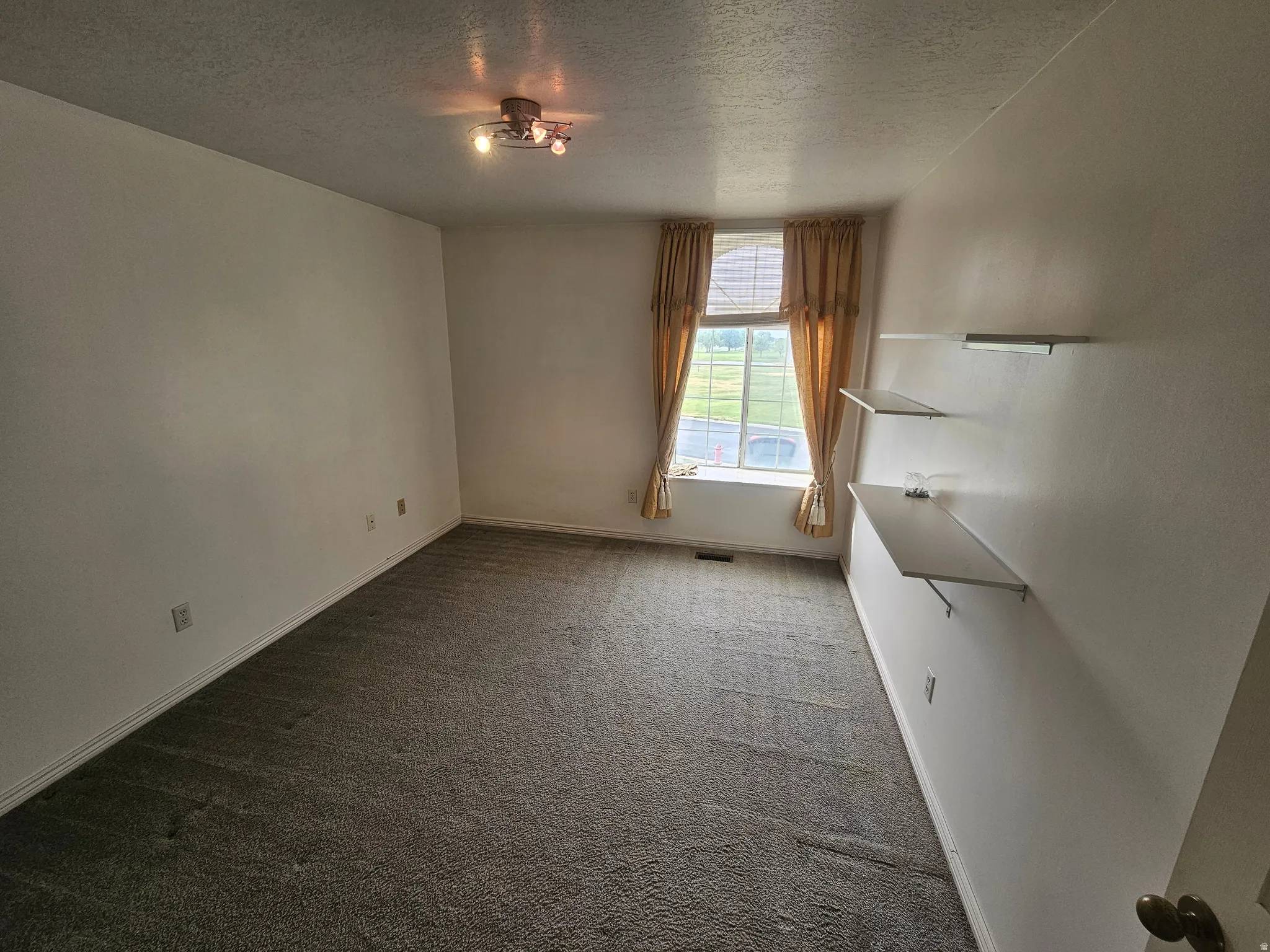 Carpeted room featuring a large window with drapes, flush-mount ceiling light fixture, and built-in wall shelves