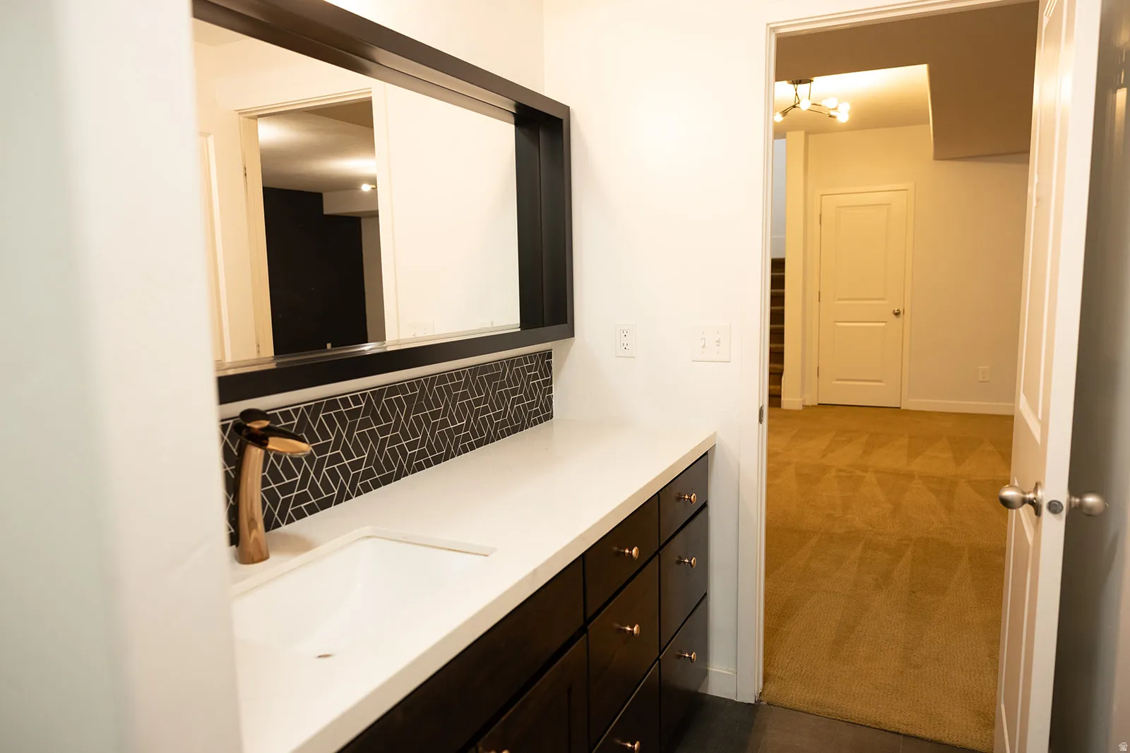 Bathroom vanity featuring a vessel sink with a unique faucet, white countertop, dark wood-finish cabinetry, geometric tile backsplash, and a framed mirror