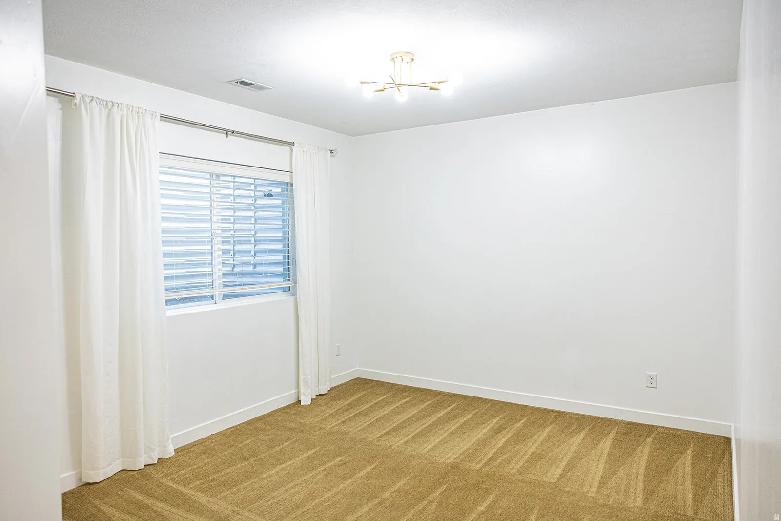 Bright room featuring a window with blinds and white drapes, white walls, and geometric-patterned carpet