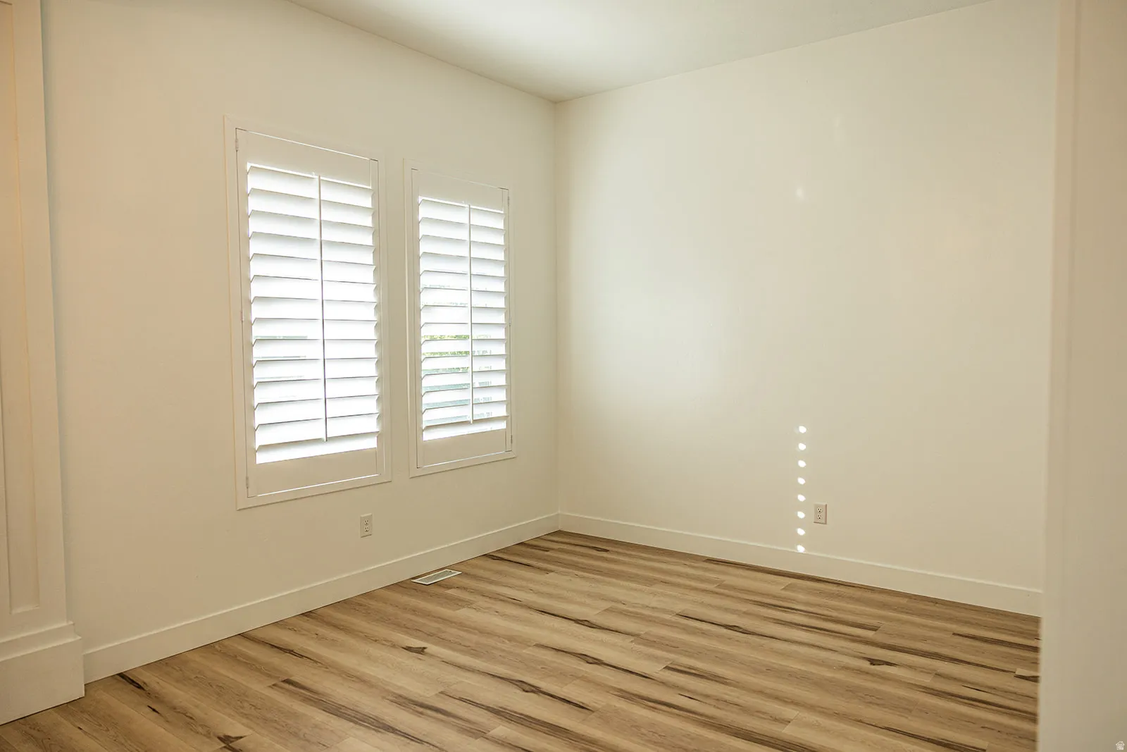 Bright interior space featuring two windows with white plantation shutters, light wood-finish flooring, white baseboards, and neutral wall paint