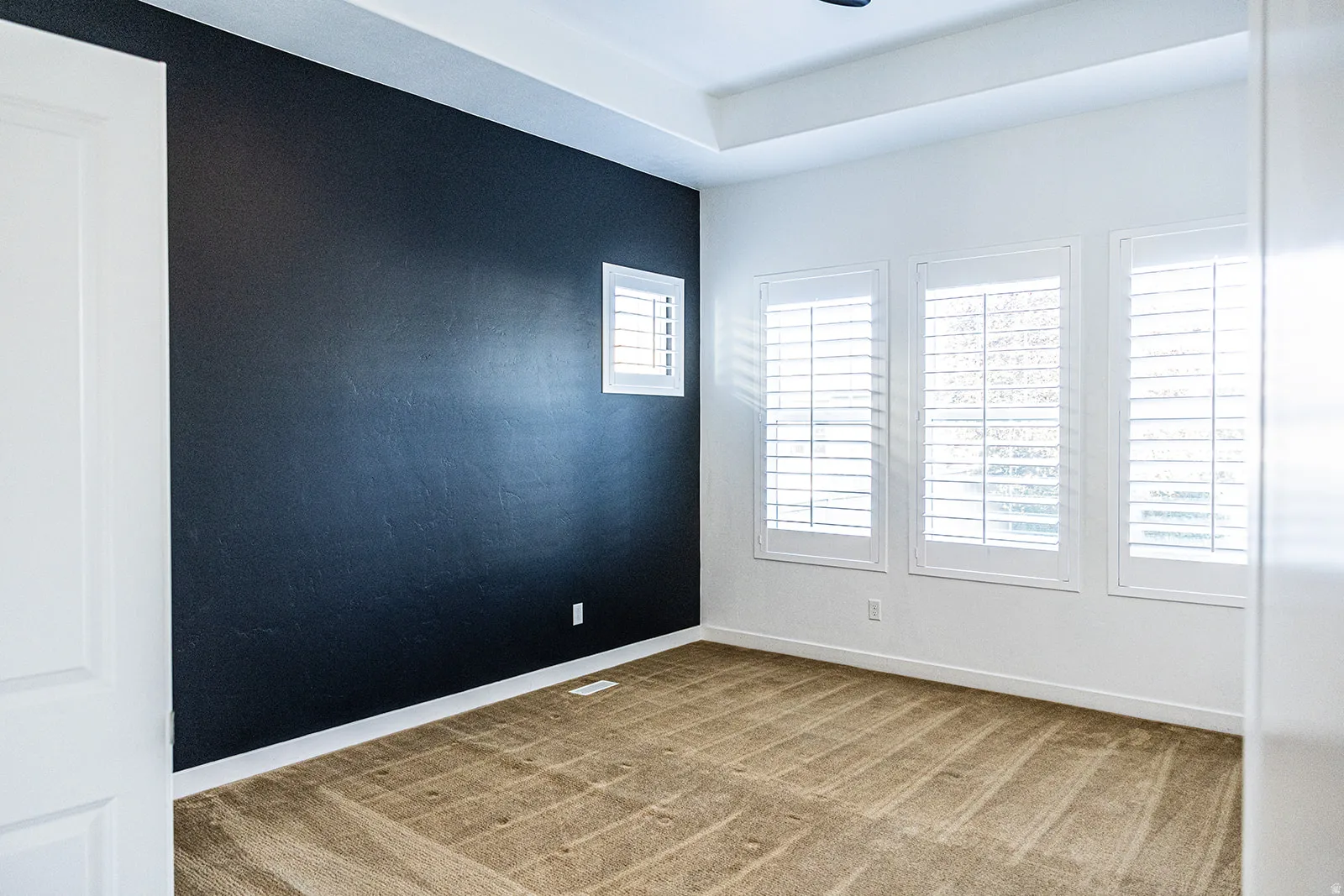 Room featuring a dark accent wall, three windows with plantation shutters, light-toned carpeting, white trim, and a tray ceiling