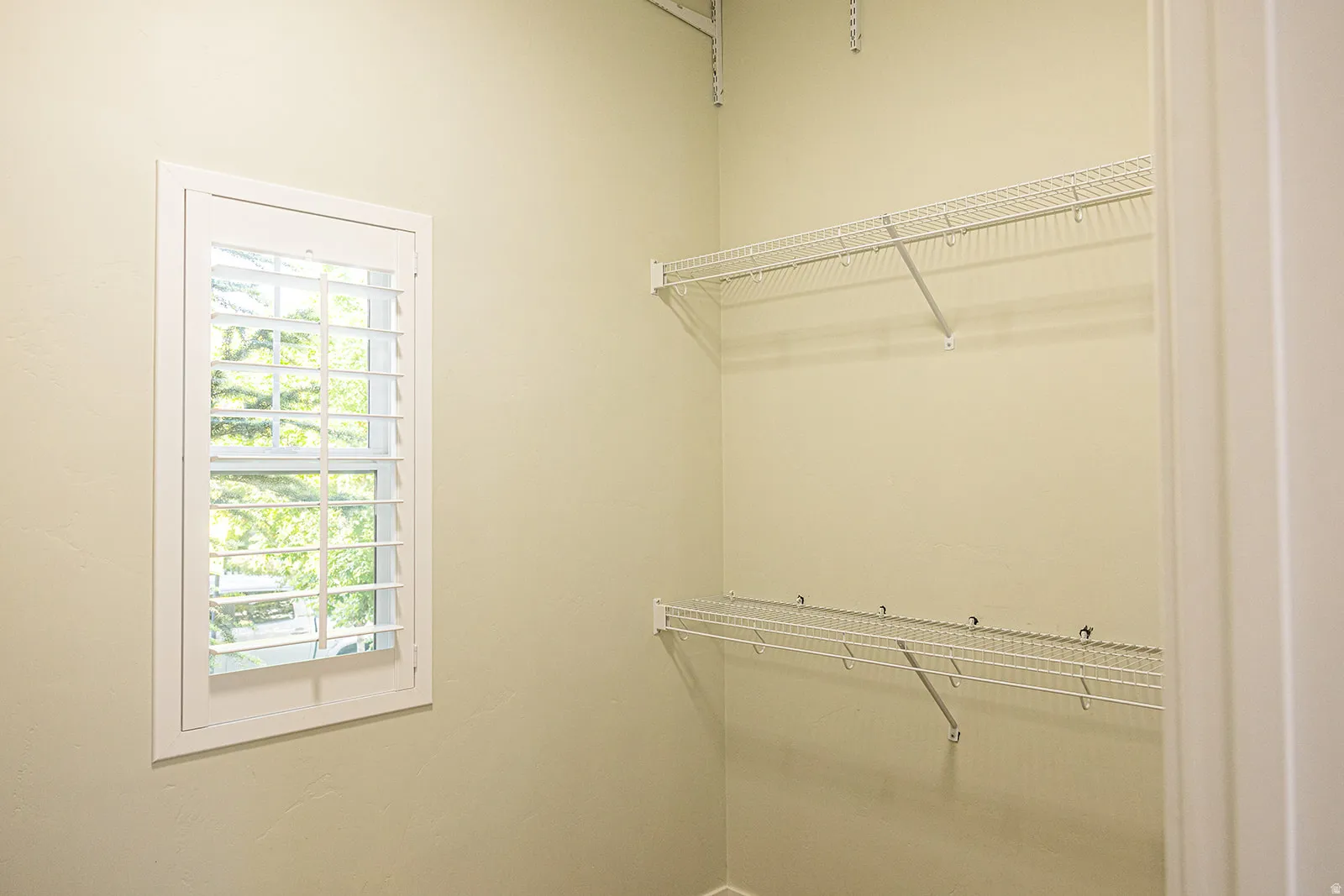 Interior space featuring a window with white plantation shutters, painted walls, and two levels of white wire shelving