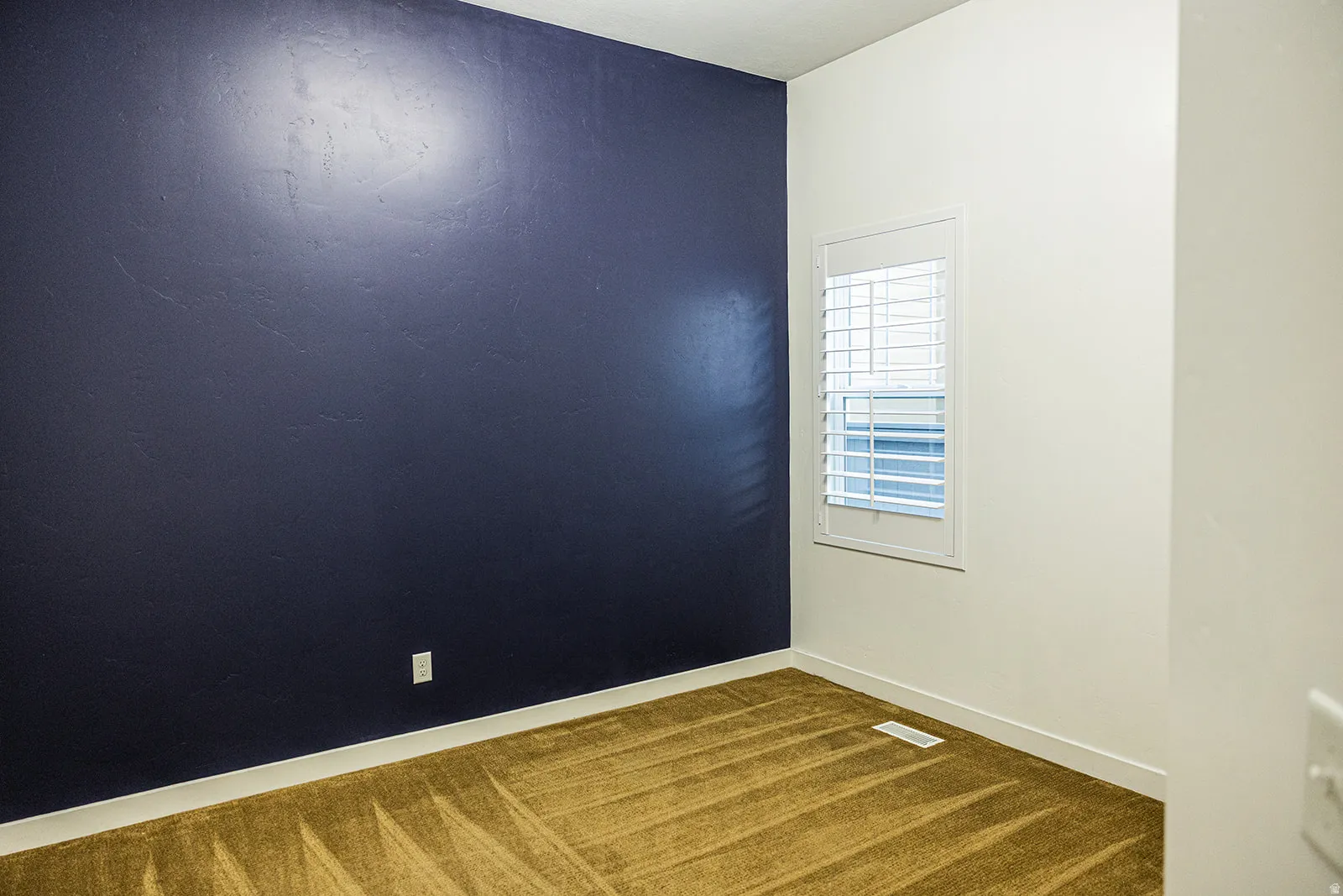 Room featuring a dark accent wall, neutral carpeting, and a window with white plantation shutters
