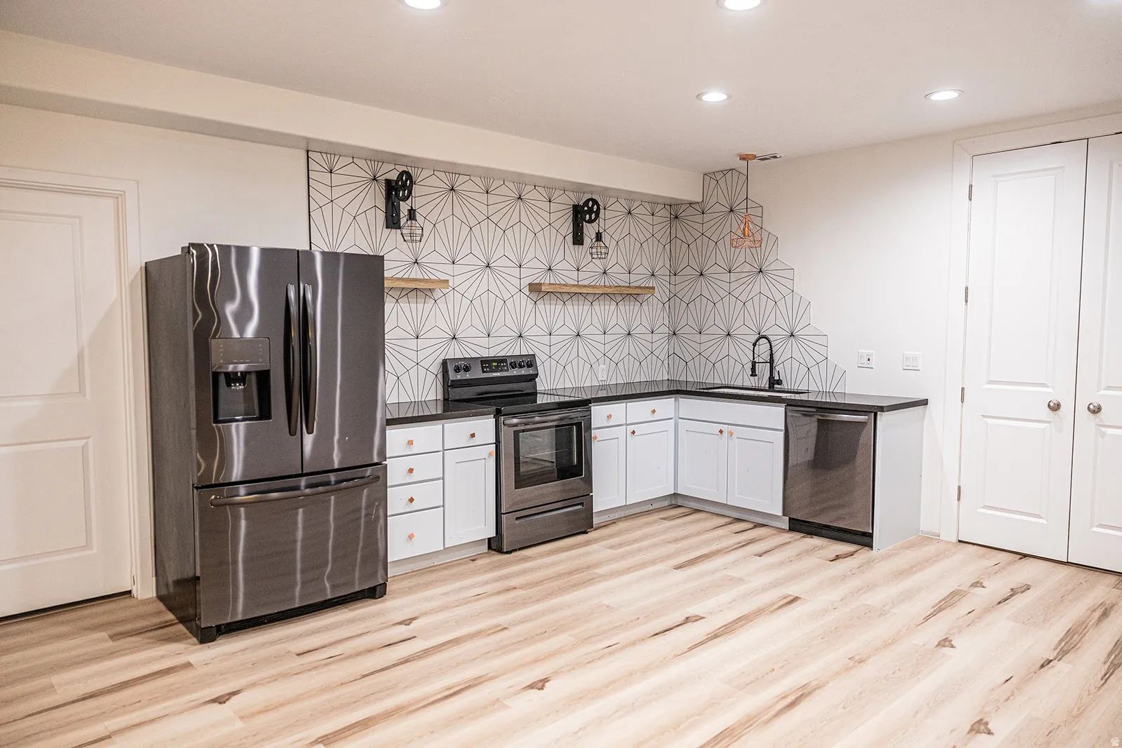 Contemporary kitchen featuring light wood-finish flooring, white cabinetry with gold-tone hardware, a geometric pattern backsplash, dark countertops, and stainless steel appliances