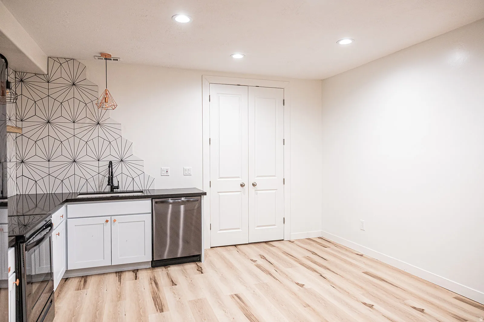 Modern kitchen area featuring white cabinetry, dark countertops, a stainless steel dishwasher, and a striking geometric tile backsplash
