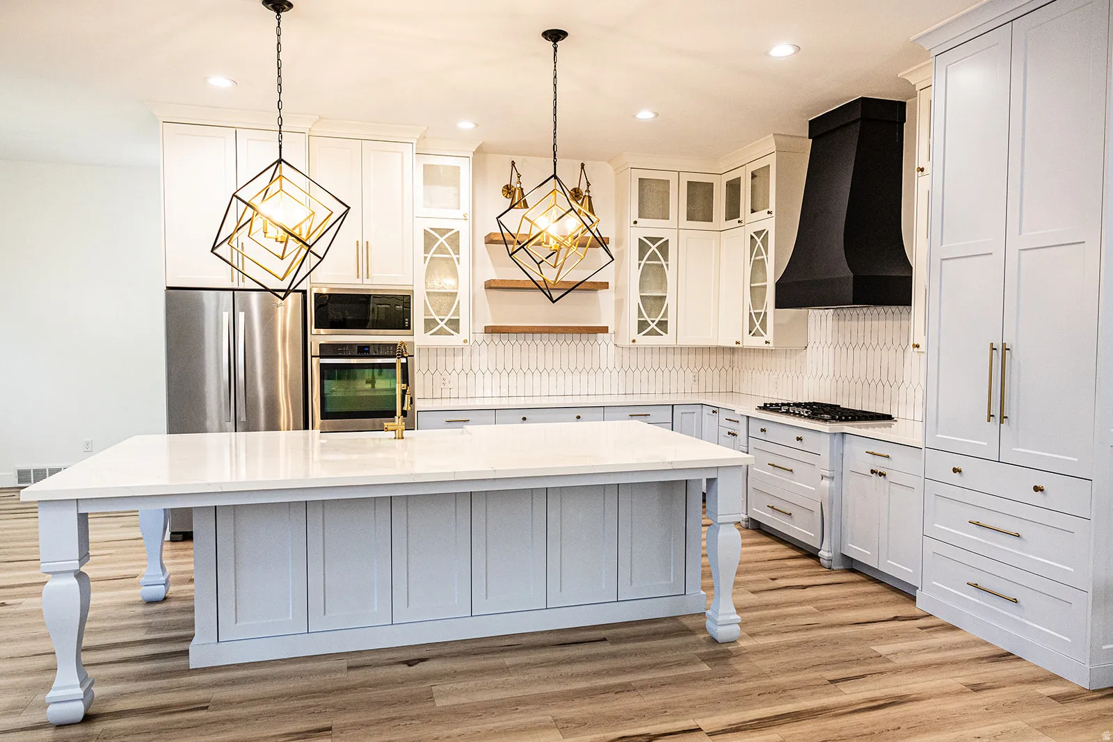 Modern kitchen featuring a large central island with a solid surface countertop, wood-finish flooring, white and light grey cabinetry, stainless steel appliances, and a black range hood