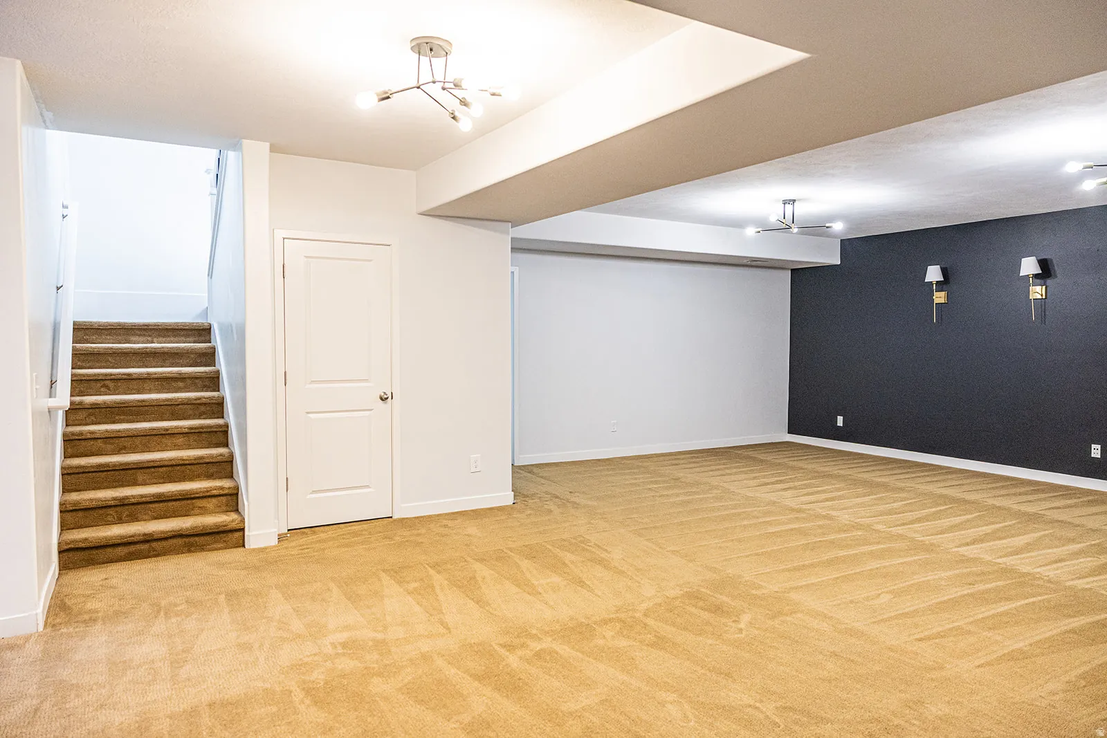 Spacious carpeted area featuring a prominent staircase with a white railing, a white interior door, a dark accent wall with two sconces, and multiple ceiling light fixtures
