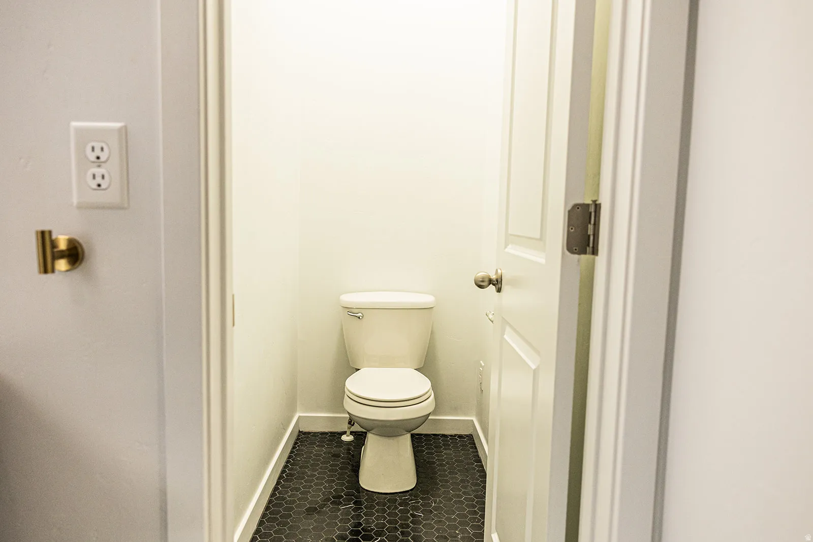 Powder room featuring a white toilet, dark hexagonal tile flooring, white baseboards, and a white paneled door with a brass-toned doorknob