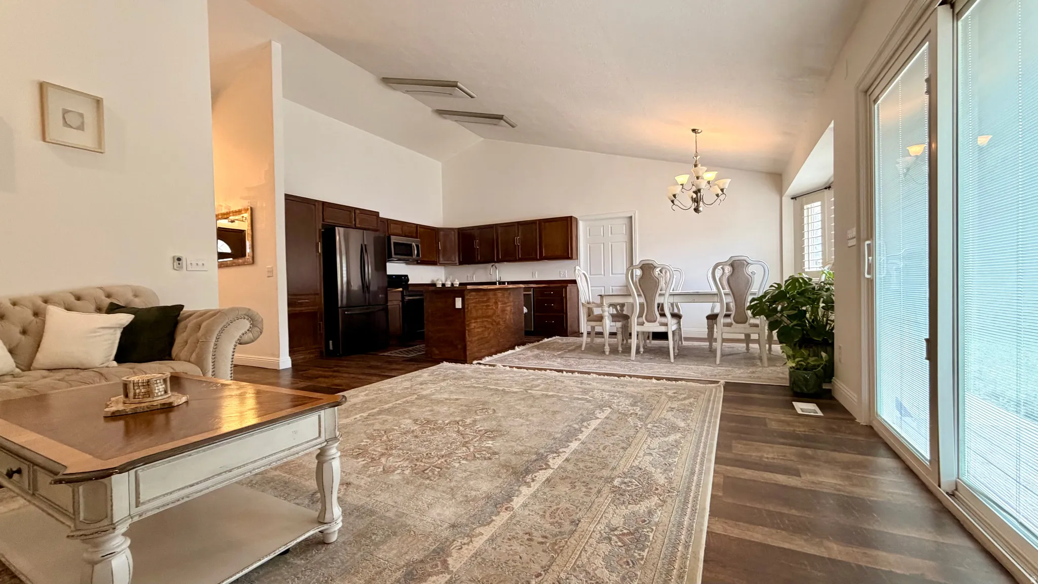 Open-concept living area featuring vaulted ceilings, wood-finish flooring, a kitchen with dark cabinetry, a dining area with chandelier lighting, and a sliding glass door