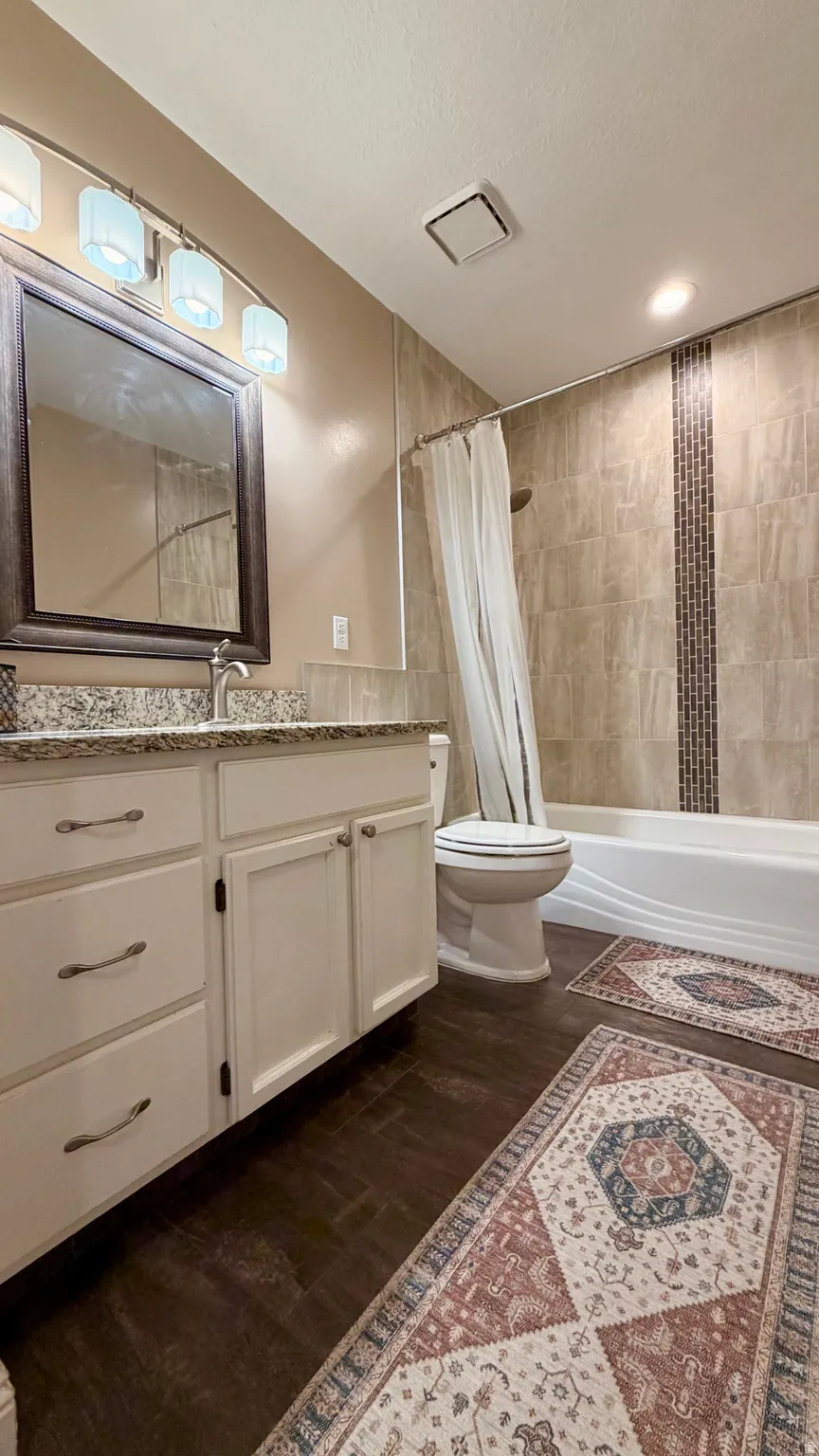 Bathroom featuring a white vanity with a granite-style countertop, dark wood-finish flooring, a white porcelain toilet, and a tiled shower-bath combination with a decorative mosaic accent strip