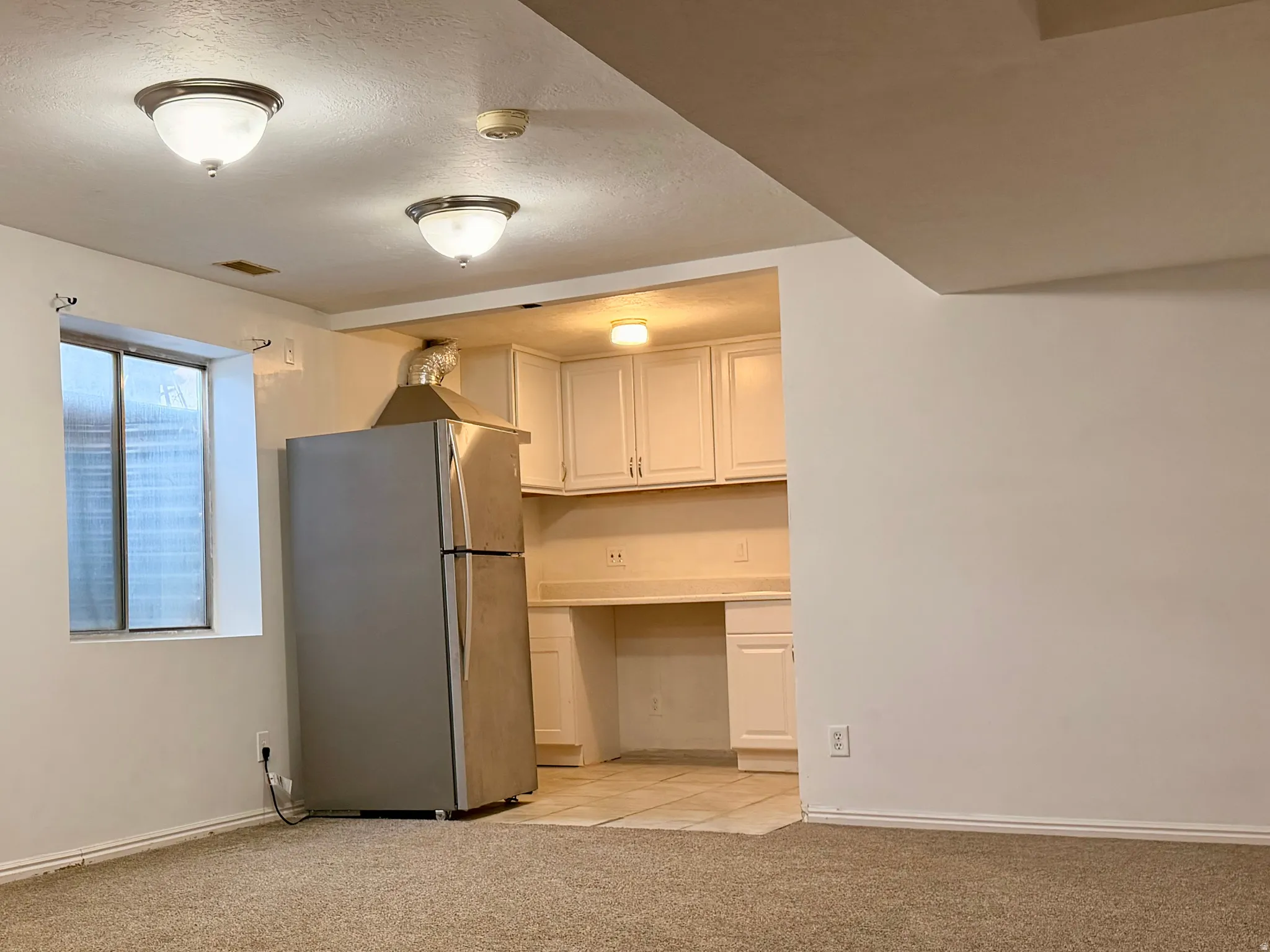 Carpeted living space opening to a kitchen area featuring white cabinetry, light countertops, and a stainless steel refrigerator