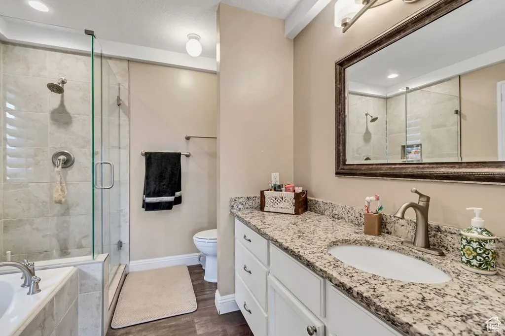 Bathroom featuring a spacious walk-in shower with glass enclosure and tiled surround, a separate soaking tub with tiled surround, a single vanity with a stone countertop, a framed mirror, and wood-finish flooring