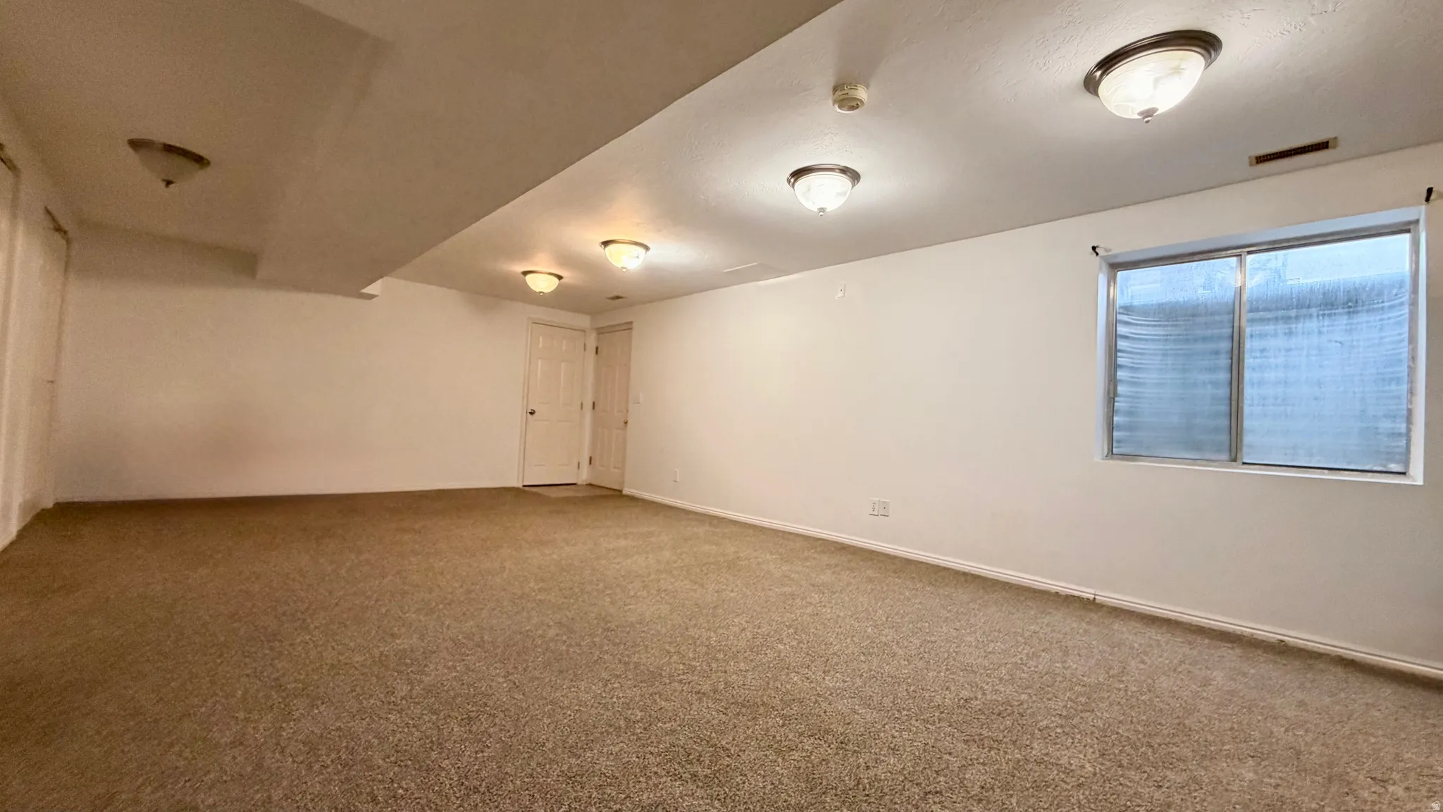 Spacious room featuring neutral carpeting, white walls, multiple flush-mount ceiling lights, and a window with blinds