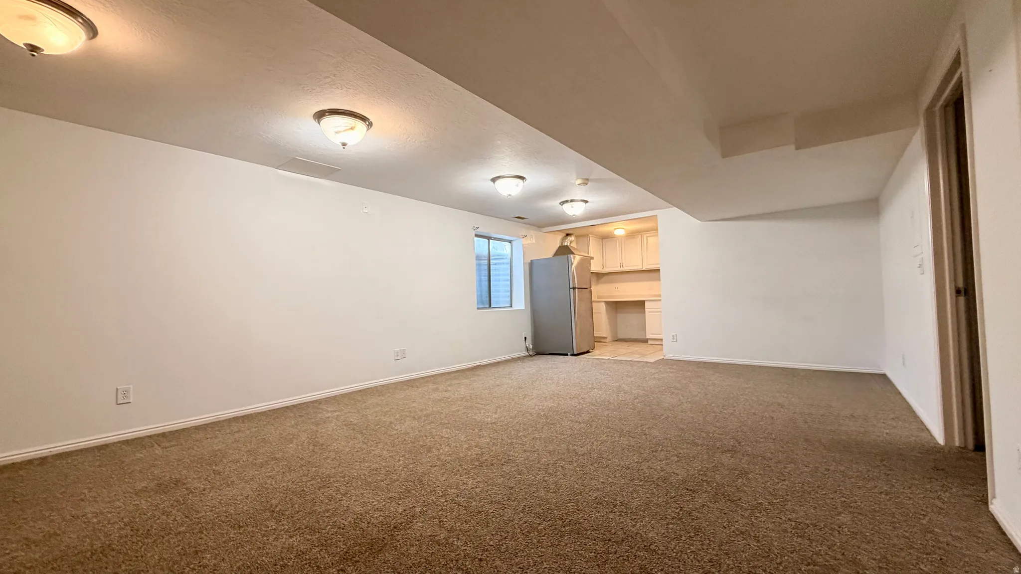 Spacious carpeted area with recessed lighting, an integrated kitchenette featuring light wood cabinetry, and a window with blinds