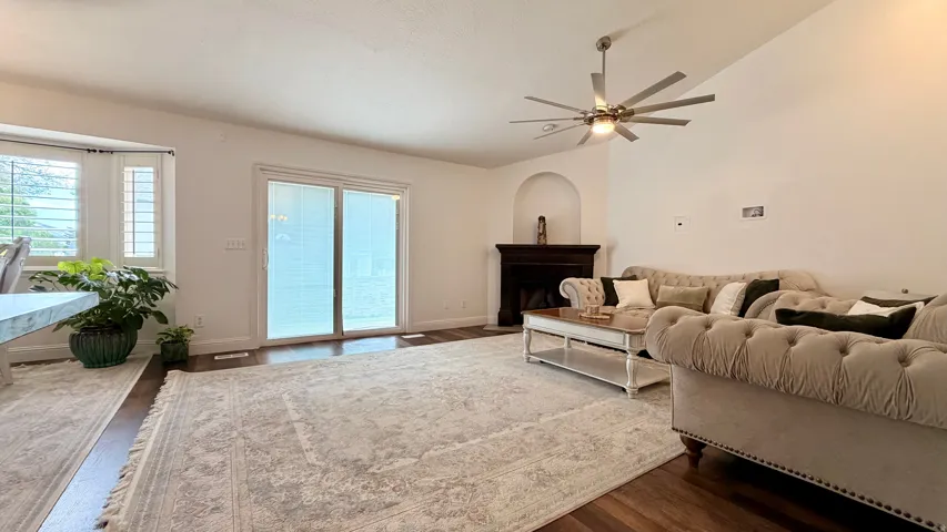 Open-concept living area featuring wood-finish flooring, a contemporary ceiling fan, and a fireplace with a dark wood mantel and arched alcove