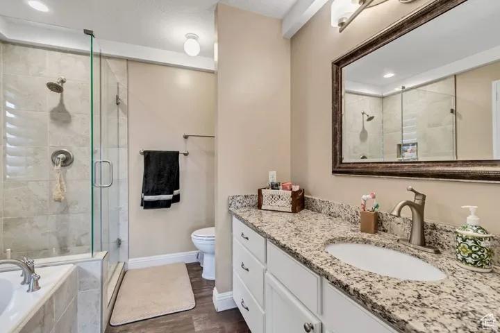 Bathroom featuring a spacious walk-in shower with glass enclosure and tiled surround, a separate soaking tub with tiled surround, a single vanity with a stone countertop, a framed mirror, and wood-finish flooring