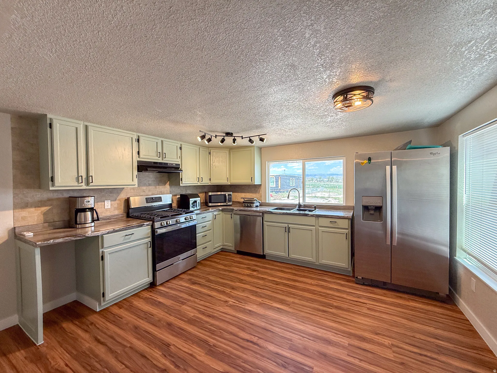 Kitchen featuring light green cabinetry, wood-finish flooring, a stainless steel gas range, stainless steel French door refrigerator, and ample counter space with a tile backsplash