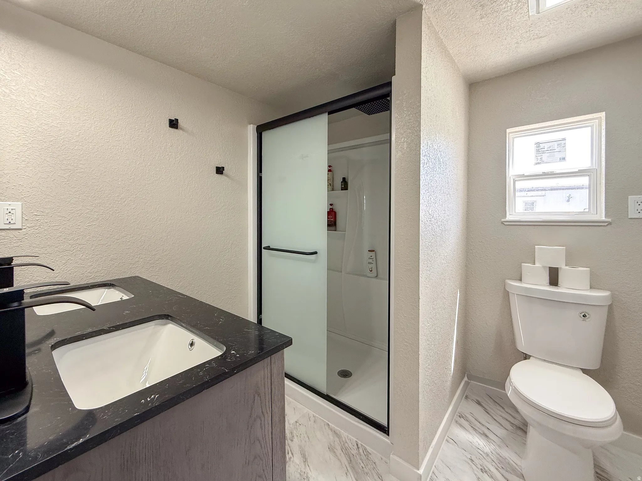 Bathroom featuring a dual vanity with a dark countertop and black fixtures, a standing shower with a frosted glass door, a toilet, and light-toned, textured walls