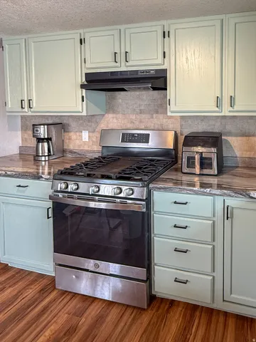 Kitchen featuring light green cabinetry with black hardware, a stainless steel range, a complementary stone-look countertop, a tiled backsplash, and wood-finish flooring