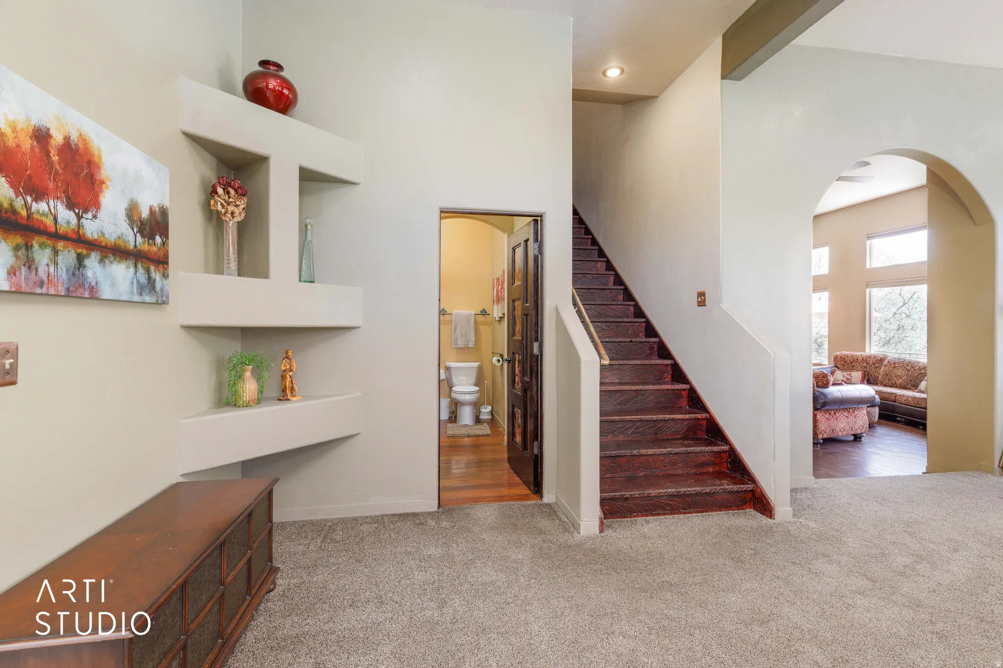 Carpeted entry area featuring a rich wood-finish staircase, built-in shelving, an arched doorway, and a visible powder room with wood-finish flooring